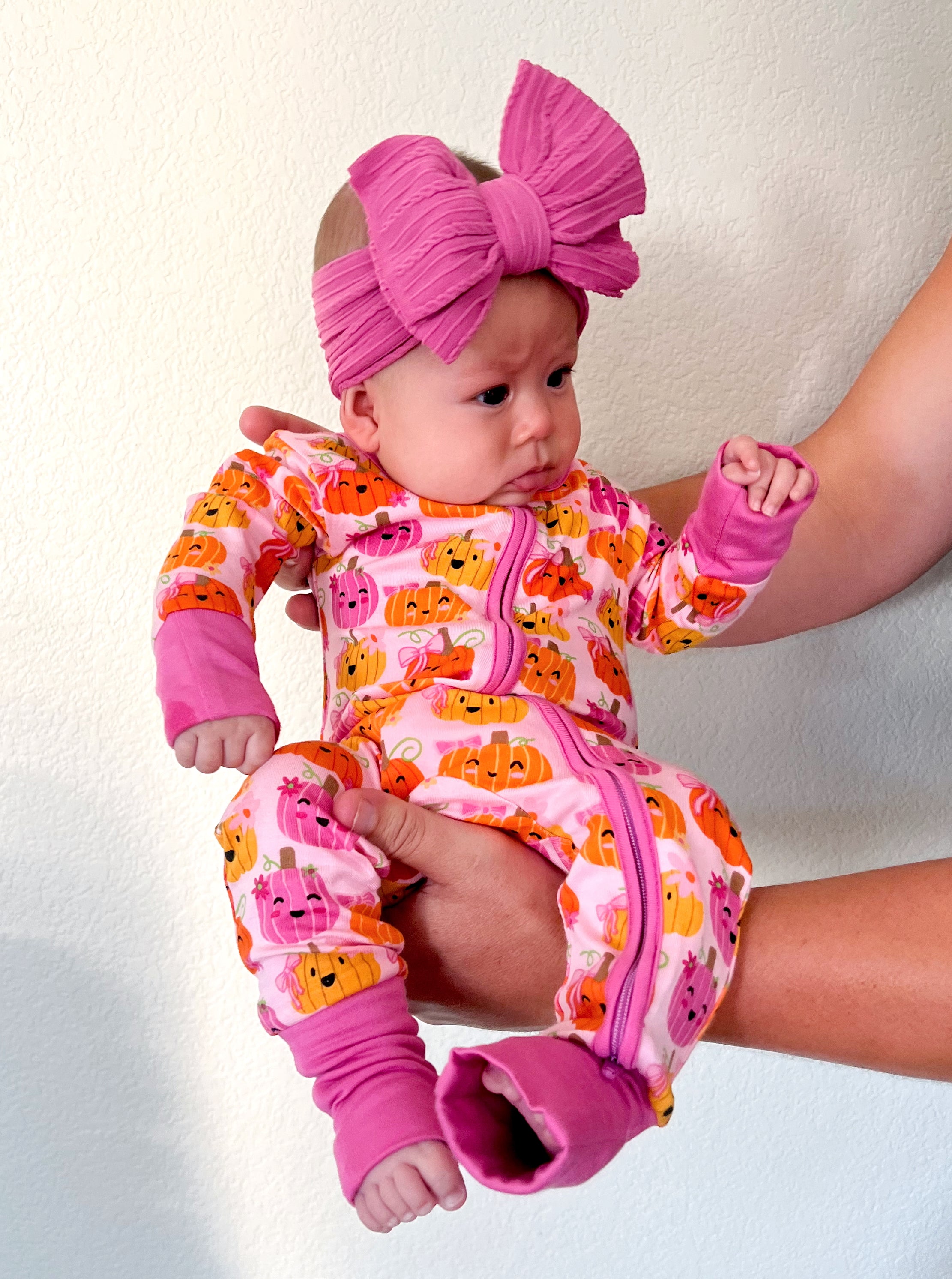 Baby in pumpkin-patterned pajamas, held by an adult, wearing a large pink headband.