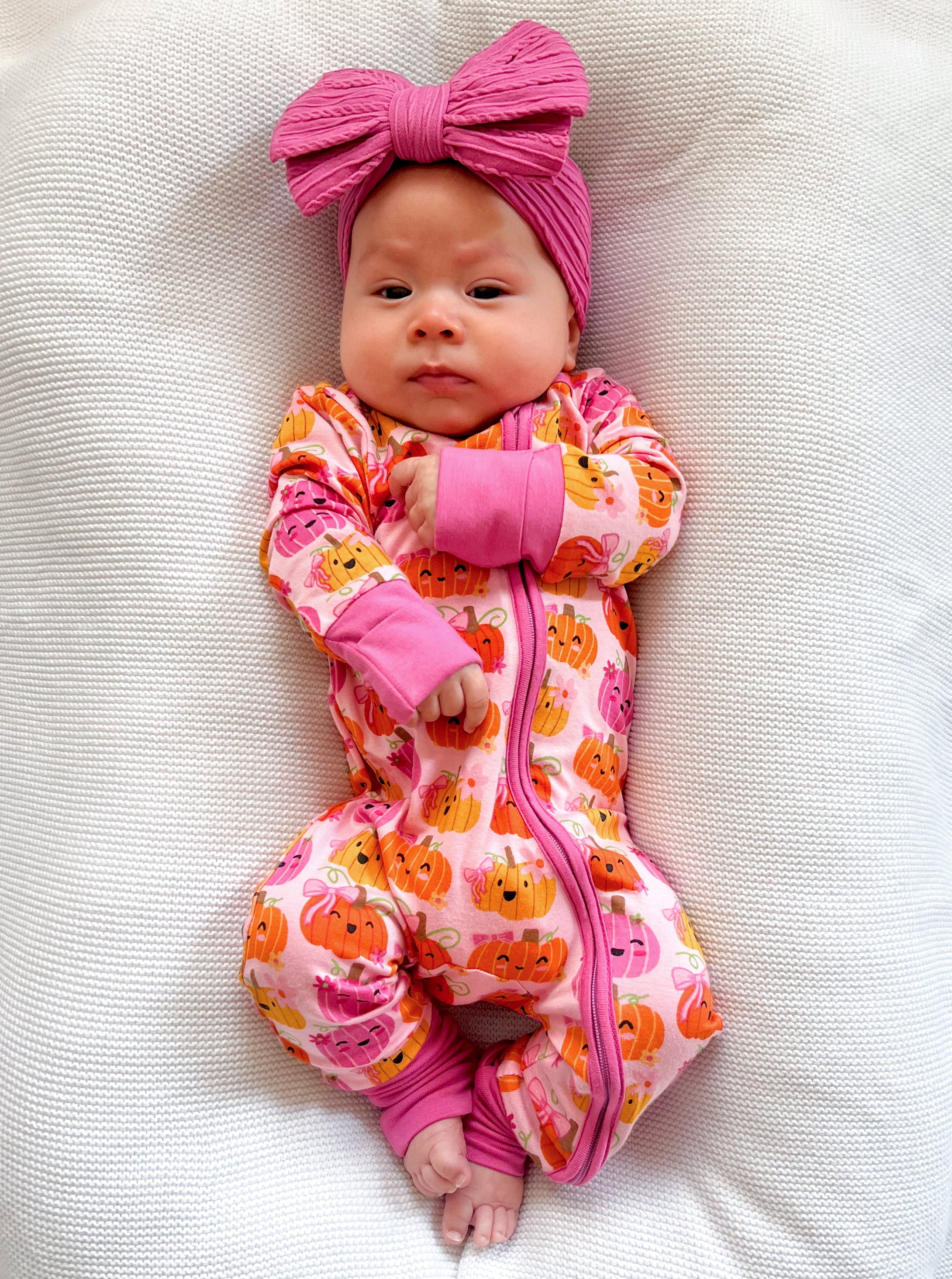 Baby in a pink pumpkin-patterned onesie and a large pink bow, lying on a textured white blanket.