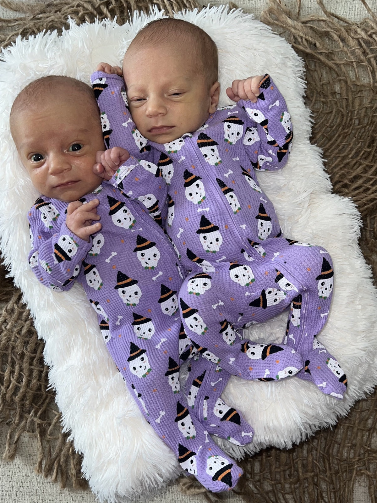 Two newborns in matching purple pajamas with ghost and skeleton patterns, resting on a fluffy white blanket.