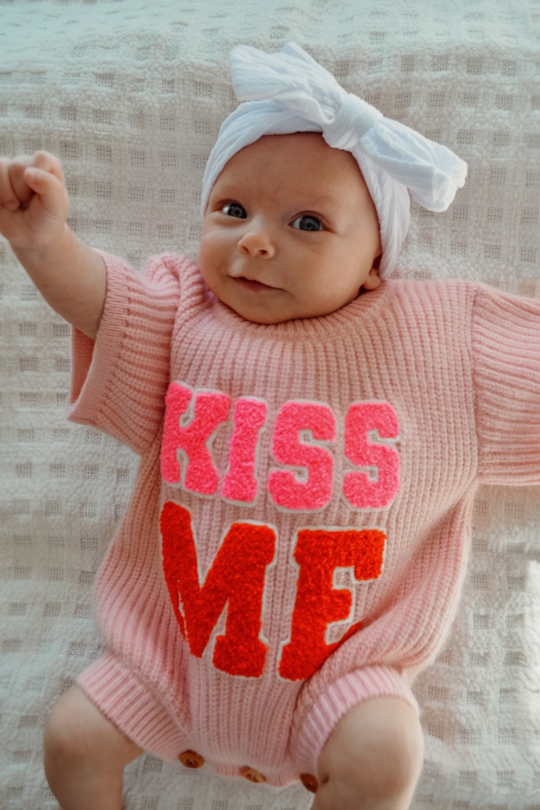 Smiling baby in a pink sweater with "KISS ME" text and a white bow headband, making a playful gesture.