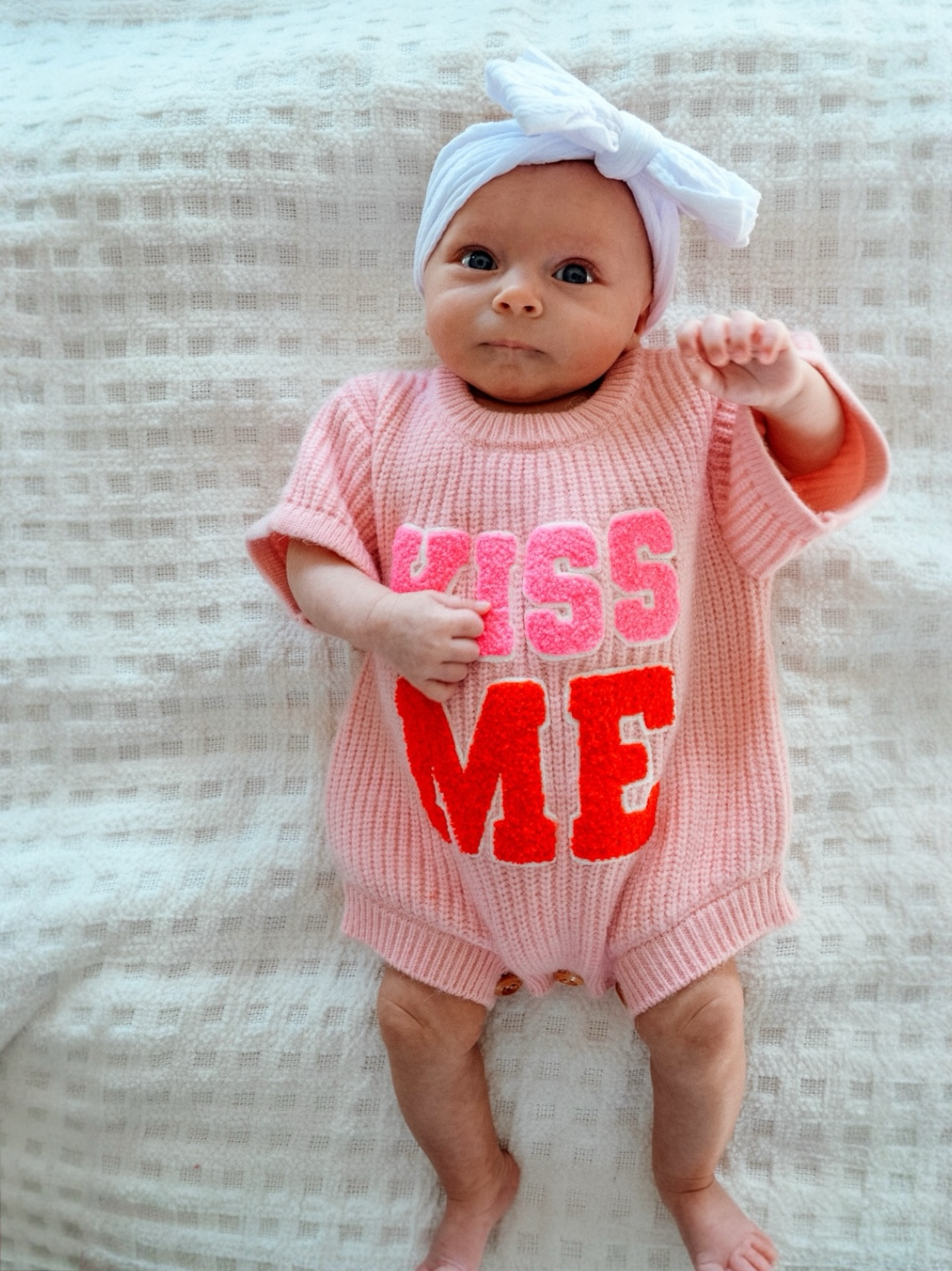 Infant in a pink outfit with "KISS ME" text, wearing a white headband, lying on a textured blanket.