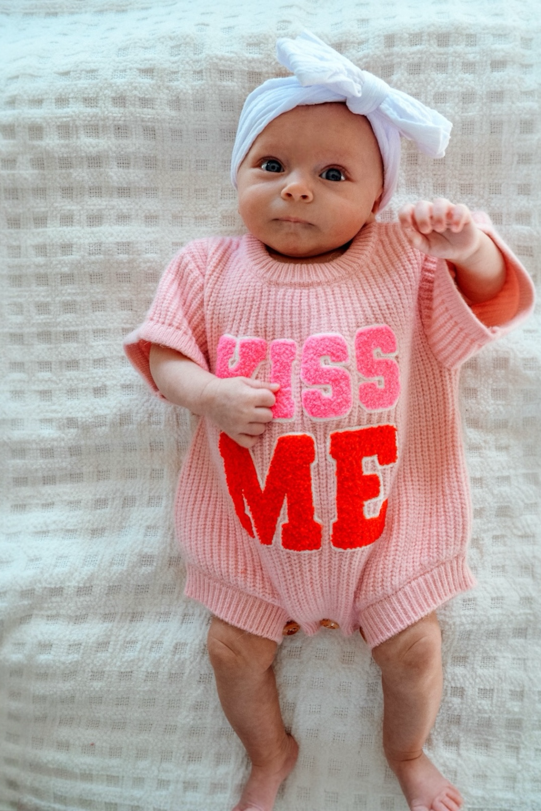 Infant in a pink outfit with "KISS ME" text, wearing a white headband, lying on a textured blanket.