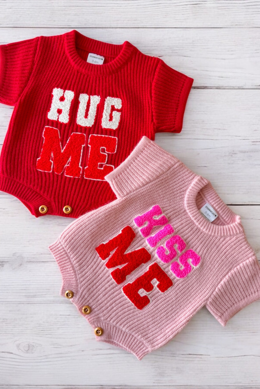 Two baby rompers: red with "HUG ME" and pink with "KISS ME," set against a light wooden background.