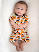 Baby lying on a white surface, wearing a pumpkin-themed outfit, looking curiously at the camera.
