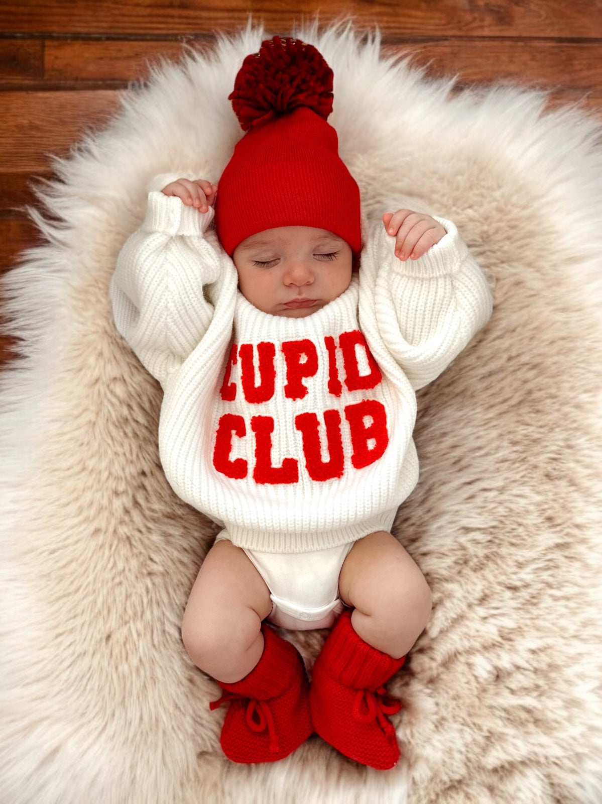 Baby wearing a cozy "Cupid Club" sweater, red hat, and booties, resting on a fluffy white blanket.