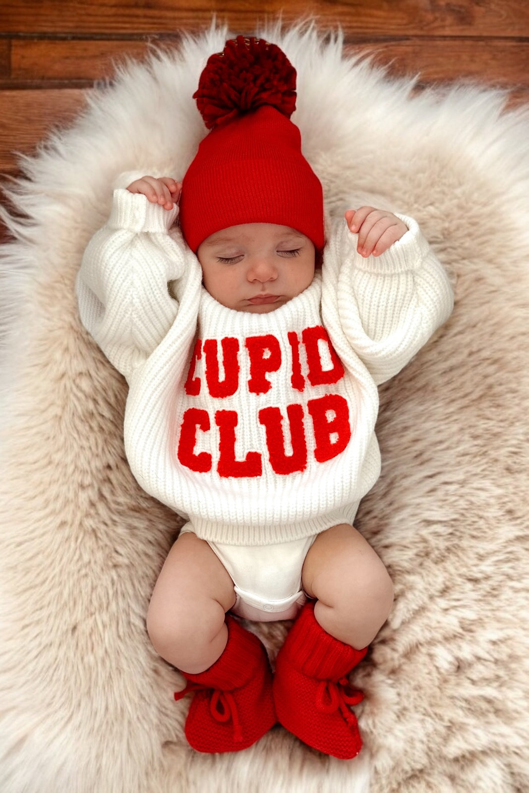 Baby wearing a cozy "Cupid Club" sweater, red hat, and booties, resting on a fluffy white blanket.