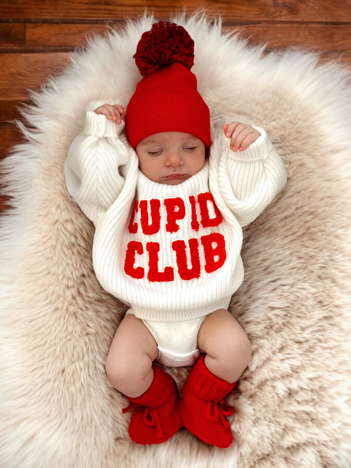 Baby wearing a cozy "Cupid Club" sweater, red hat, and booties, resting on a fluffy white blanket.