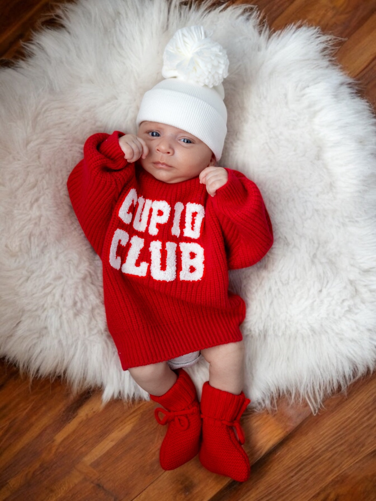 Baby in a red "Cupid Club" sweater and white hat, lying on a fluffy white rug with wooden floor beneath.