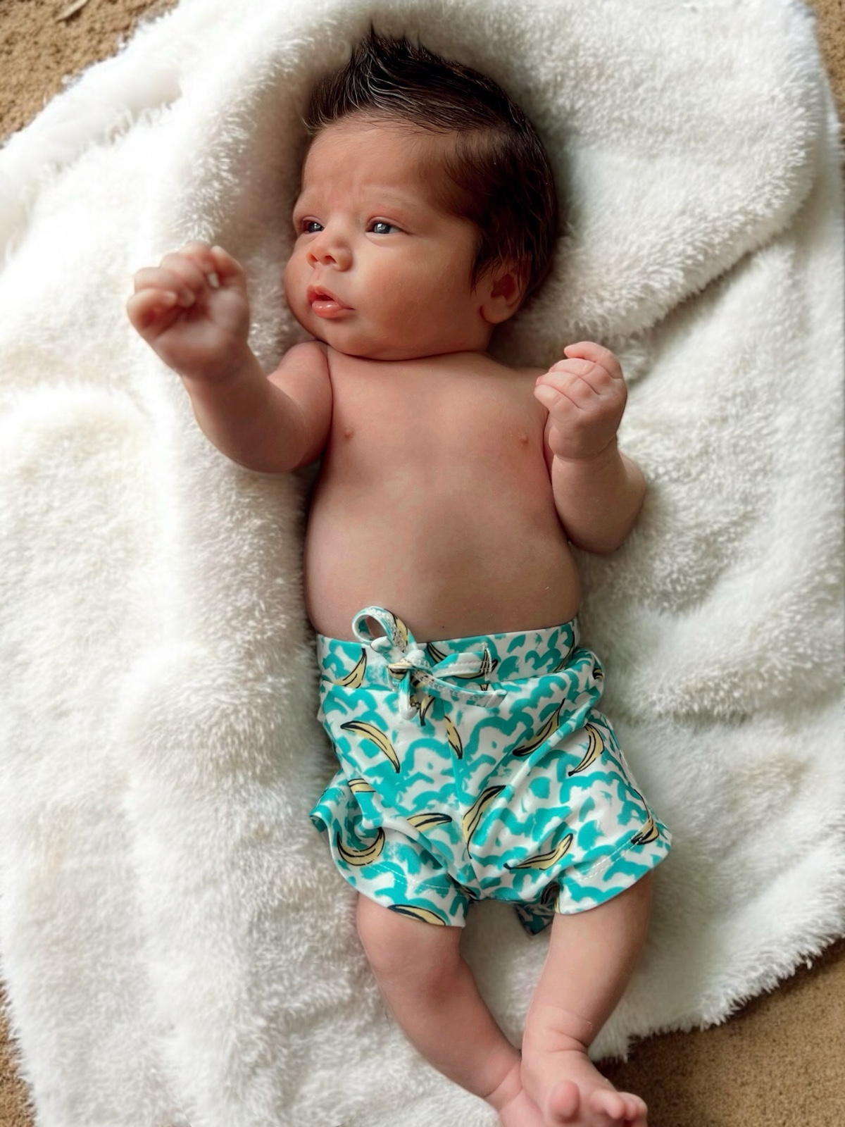 Baby with spiky hair lying on a fluffy blanket, wearing blue shorts with banana print, making a fist.