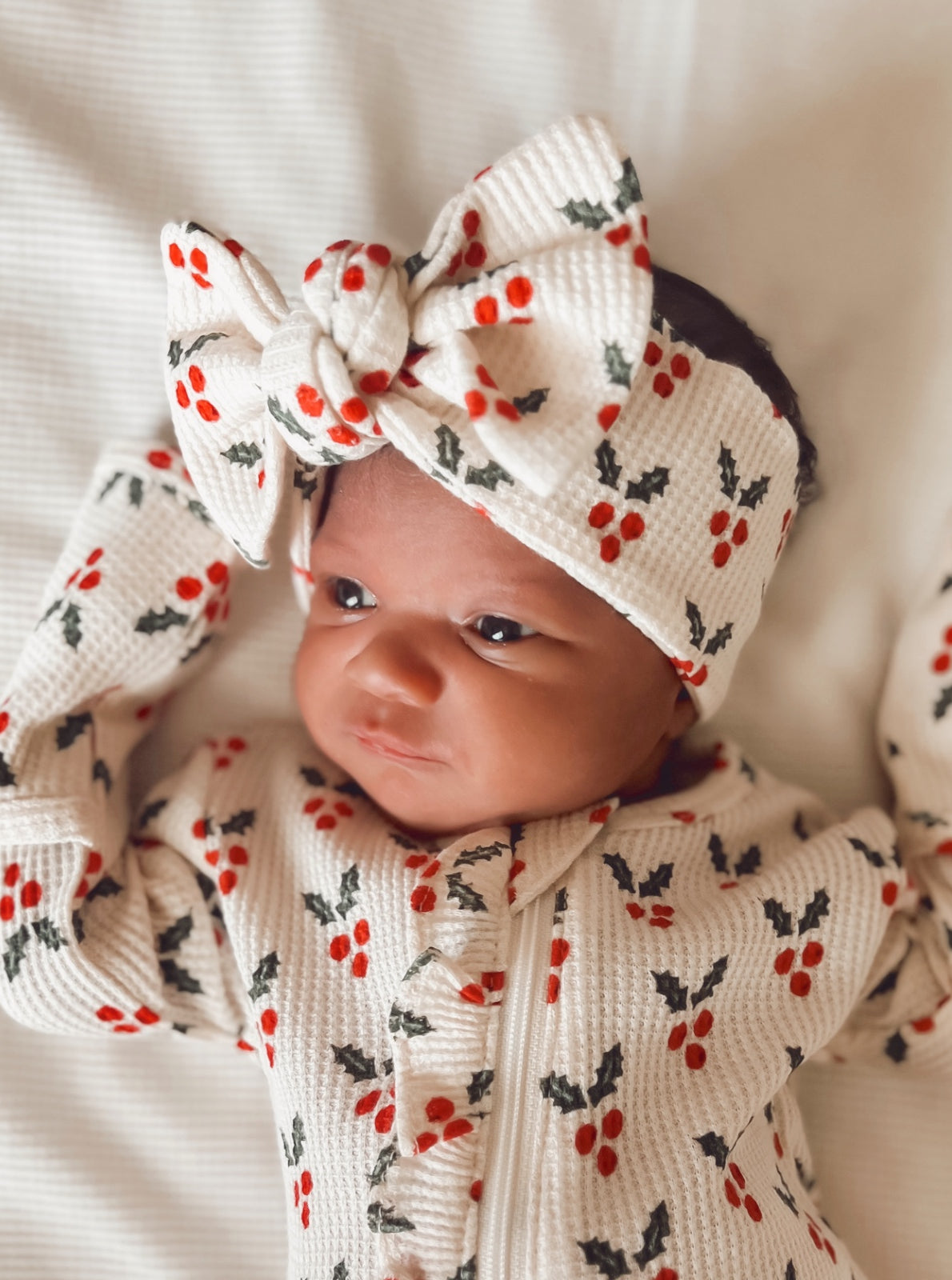 Newborn wearing a festive outfit with holly patterns and a large bow headband, looking curiously at the camera.