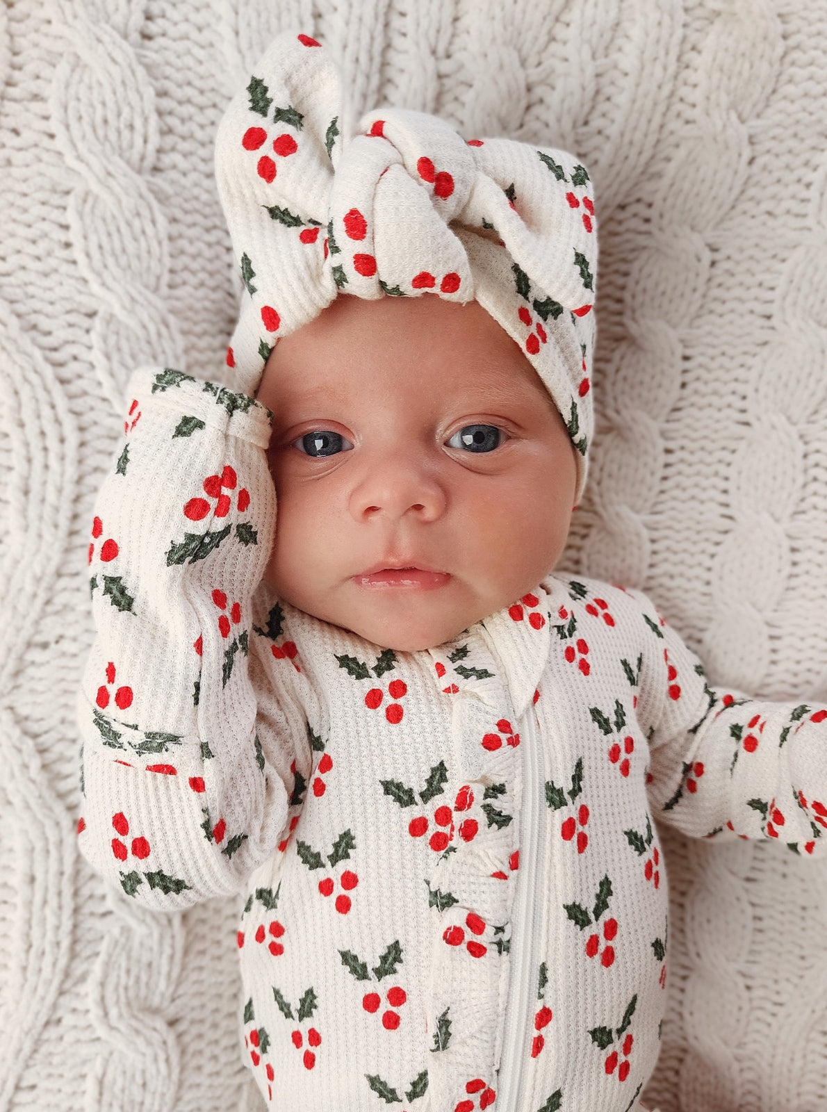 Infant wearing a holiday-themed onesie with red berries and a matching headband, resting on a textured blanket.