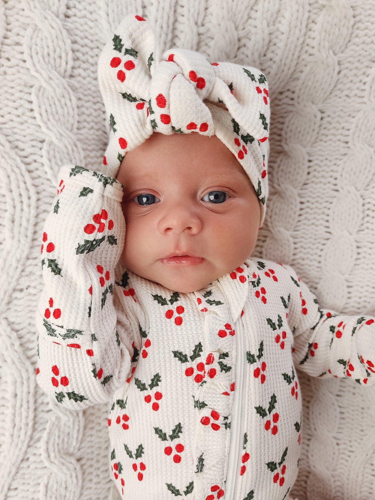 Infant wearing a holiday-themed onesie with red berries and a matching headband, resting on a textured blanket.