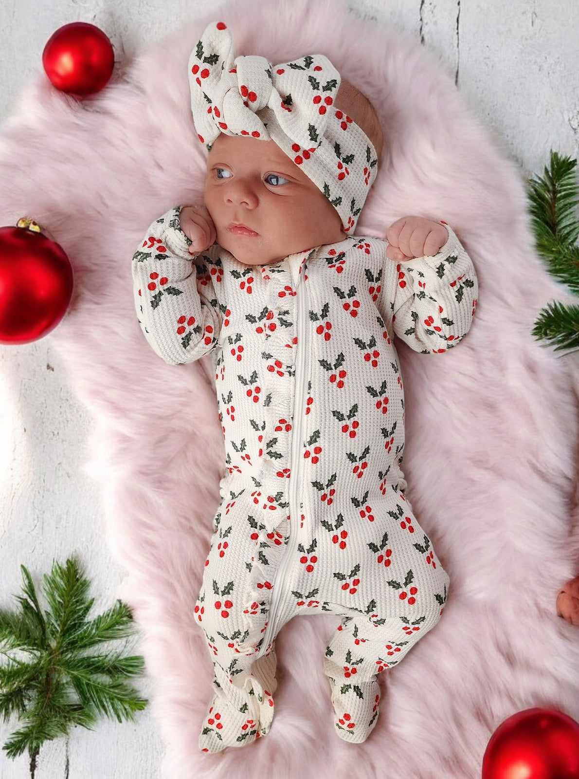 Baby in holiday-themed outfit with cherry pattern, wearing a matching headband, on fluffy blanket surrounded by ornaments.