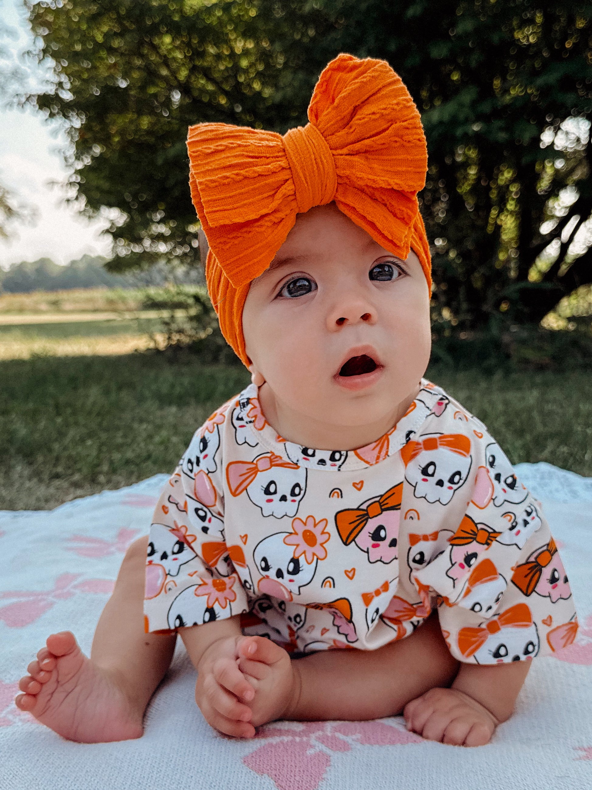 Baby sitting on a blanket outdoors, wearing a colorful skull-patterned outfit and a large orange bow headband.