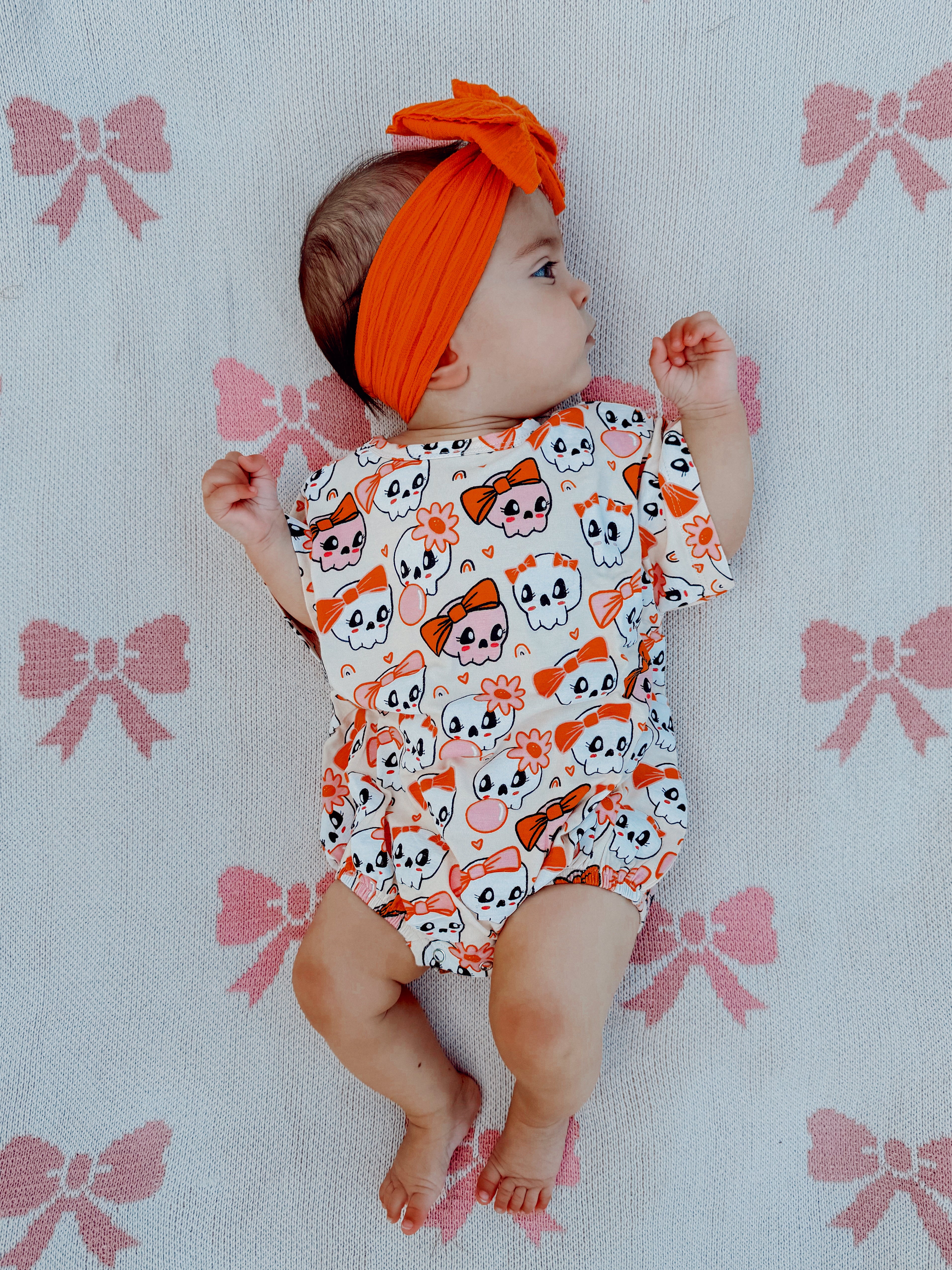 Baby lying on a patterned blanket, wearing a colorful skull print outfit and orange headband, smiling.