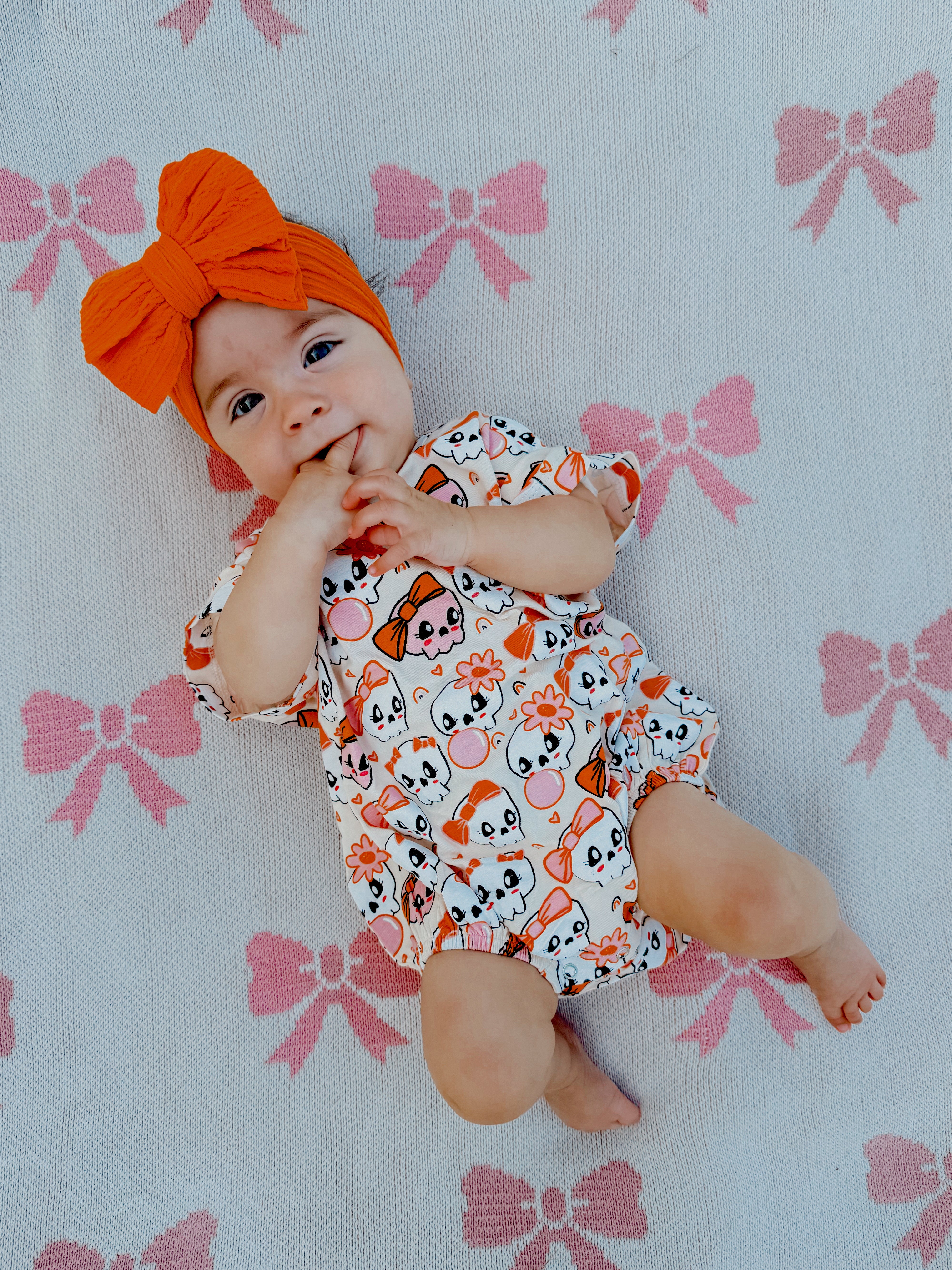 Baby with an orange bow headband, smiling on a pink bow-patterned blanket, wearing a playful animal print outfit.