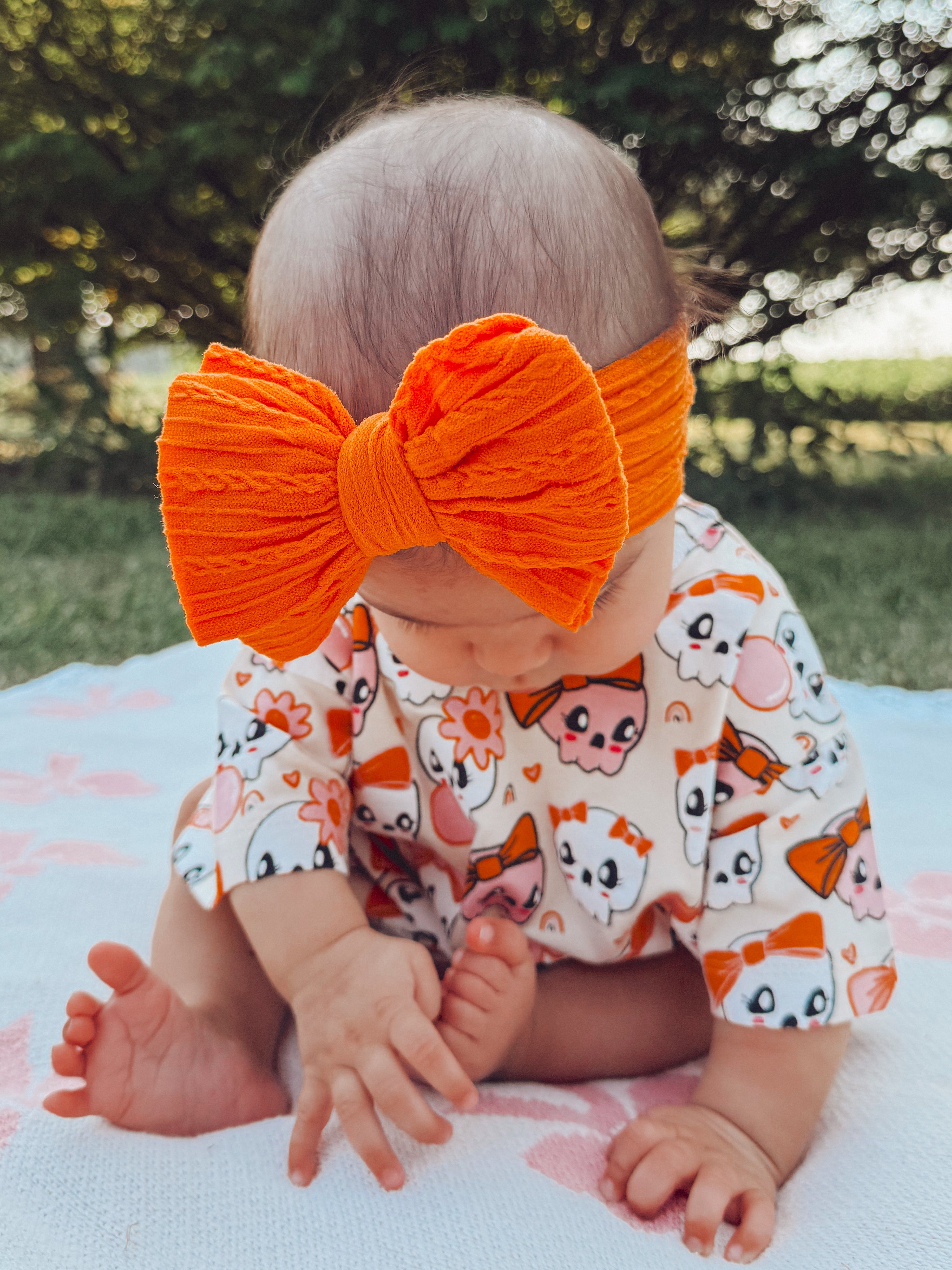 Baby wearing a large orange bow and a playful outfit, sitting on a blanket in a sunny outdoor setting.