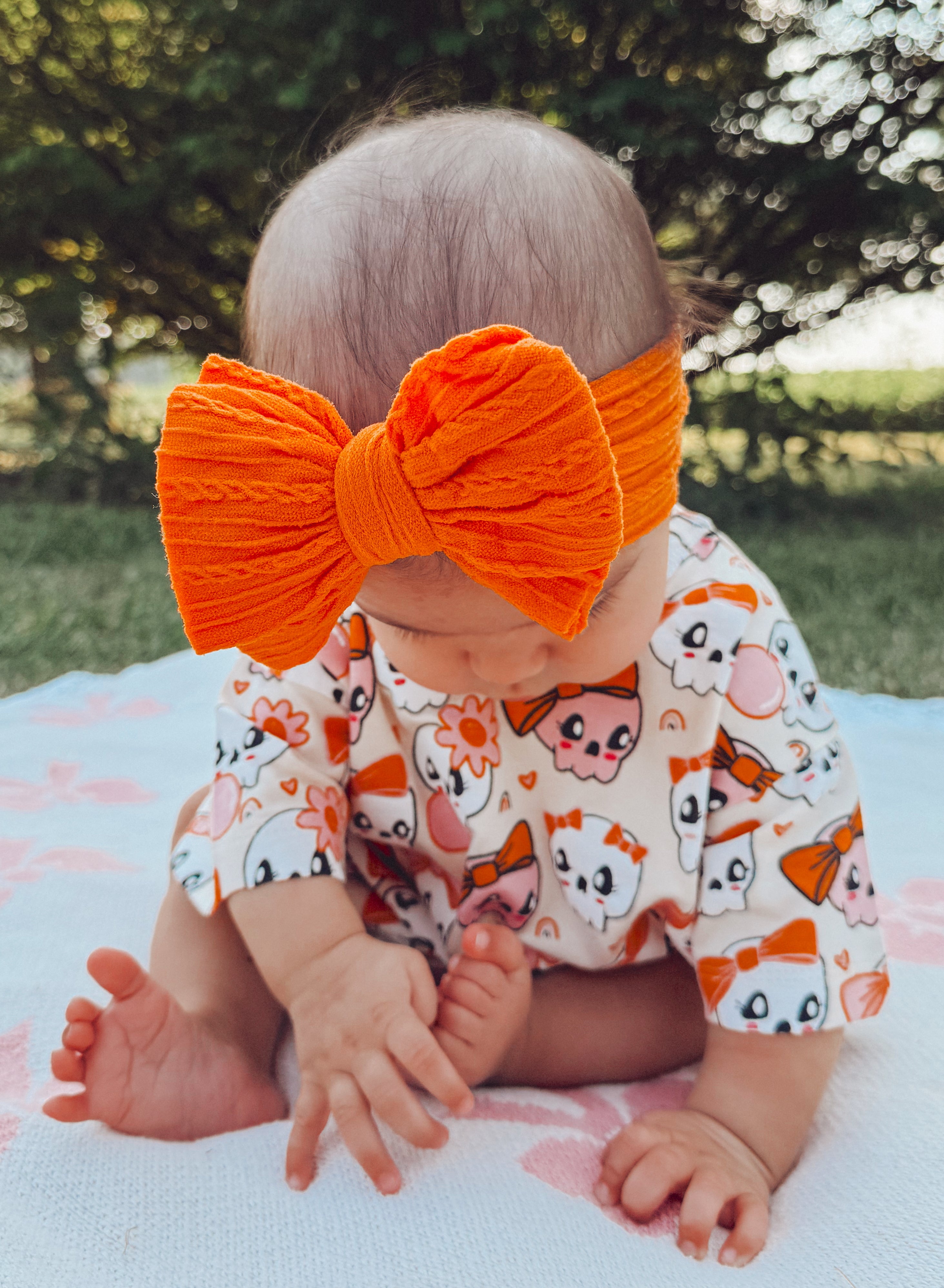 Baby wearing a large orange bow and a playful outfit, sitting on a blanket in a sunny outdoor setting.