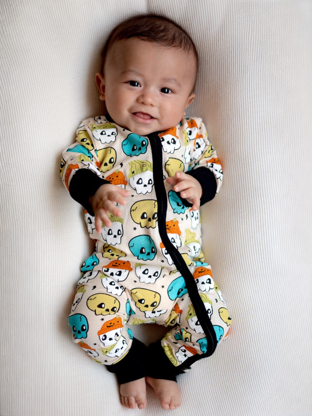 Smiling baby in colorful skull-print pajamas, sitting on a light textured surface.