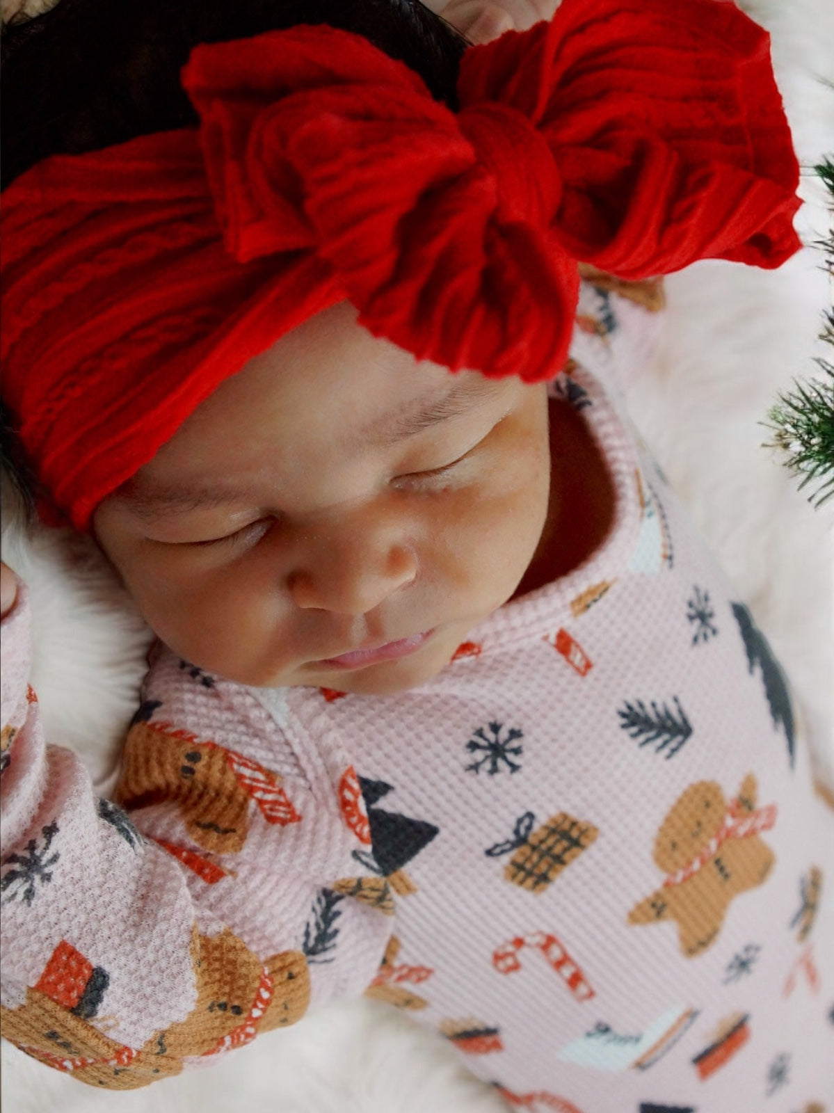 Sleeping baby in festive pajamas and a red bow headband, resting on a soft white blanket.