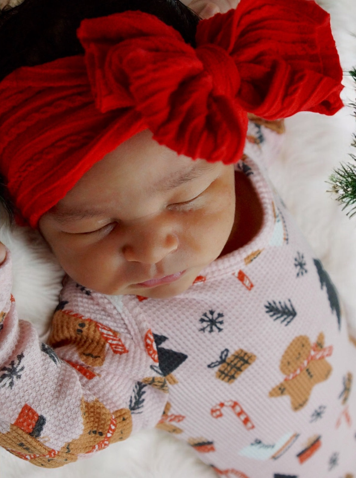 Sleeping baby in festive pajamas and a red bow headband, resting on a soft white blanket.