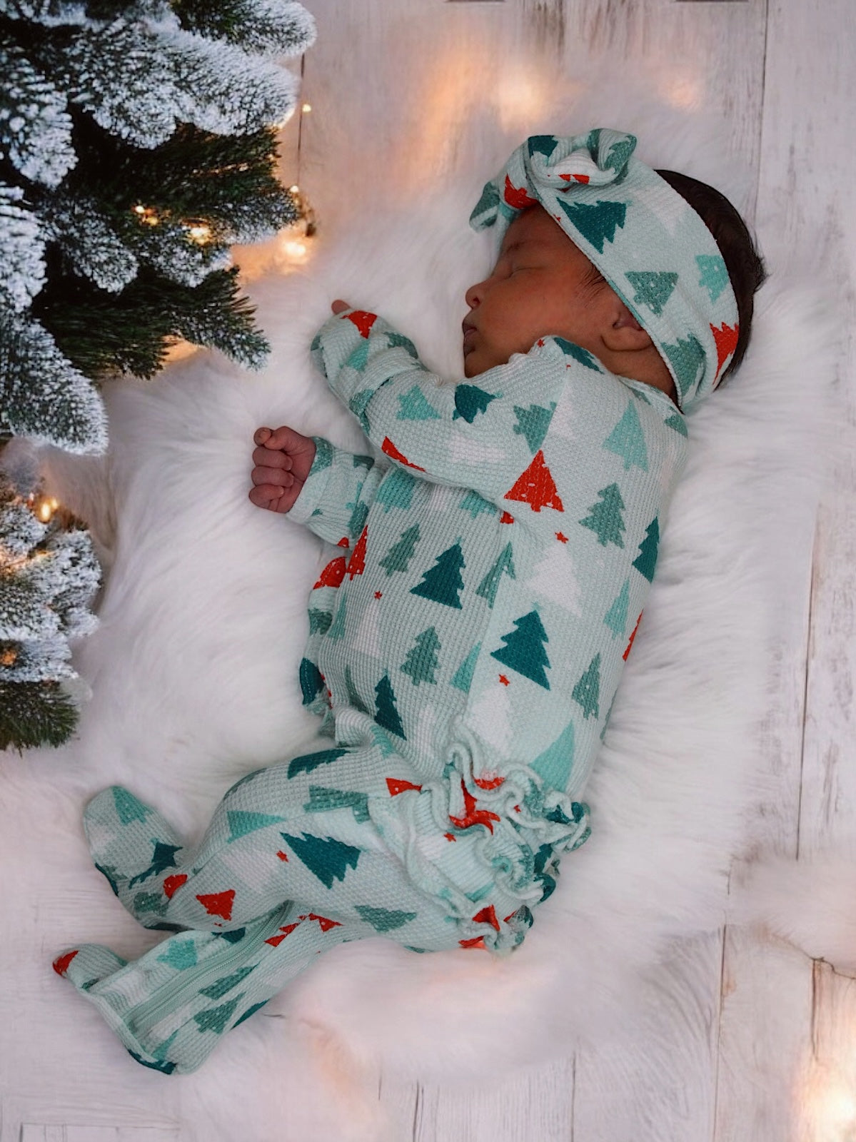 Sleeping baby in festive pajamas beside a decorated tree, on a fluffy white rug with soft lighting.