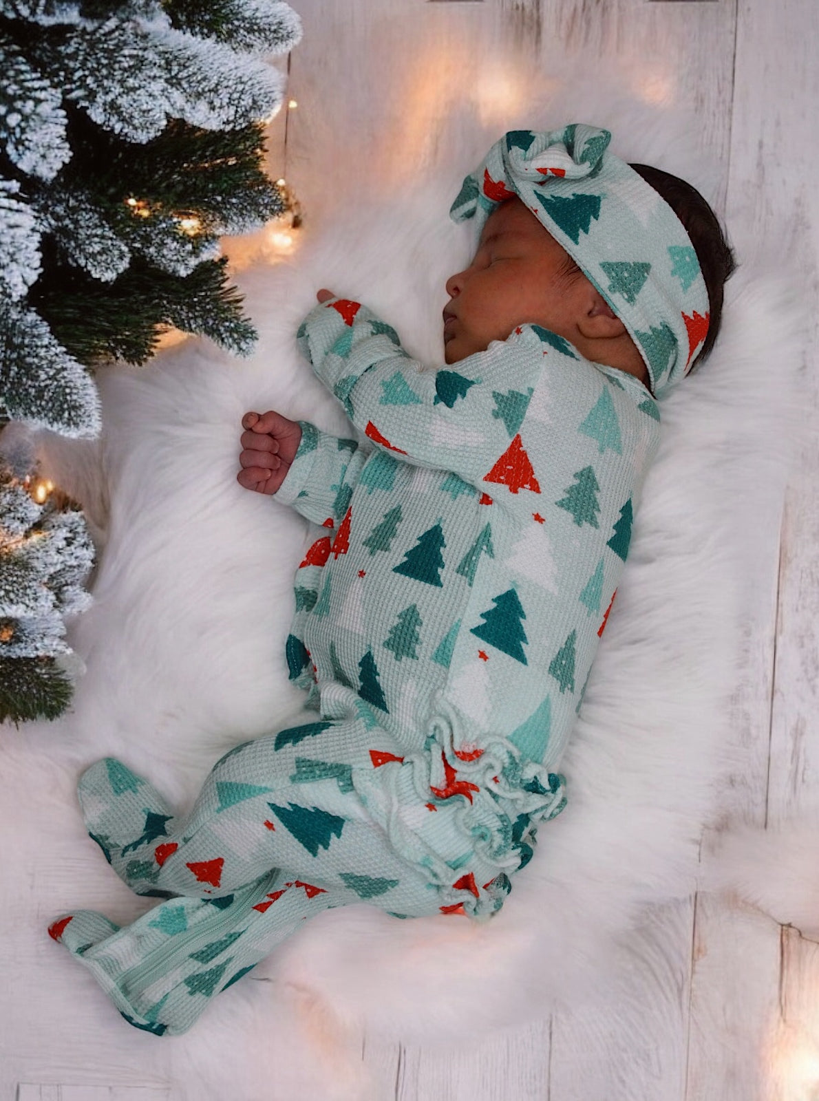 Sleeping baby in festive pajamas beside a decorated tree, on a fluffy white rug with soft lighting.