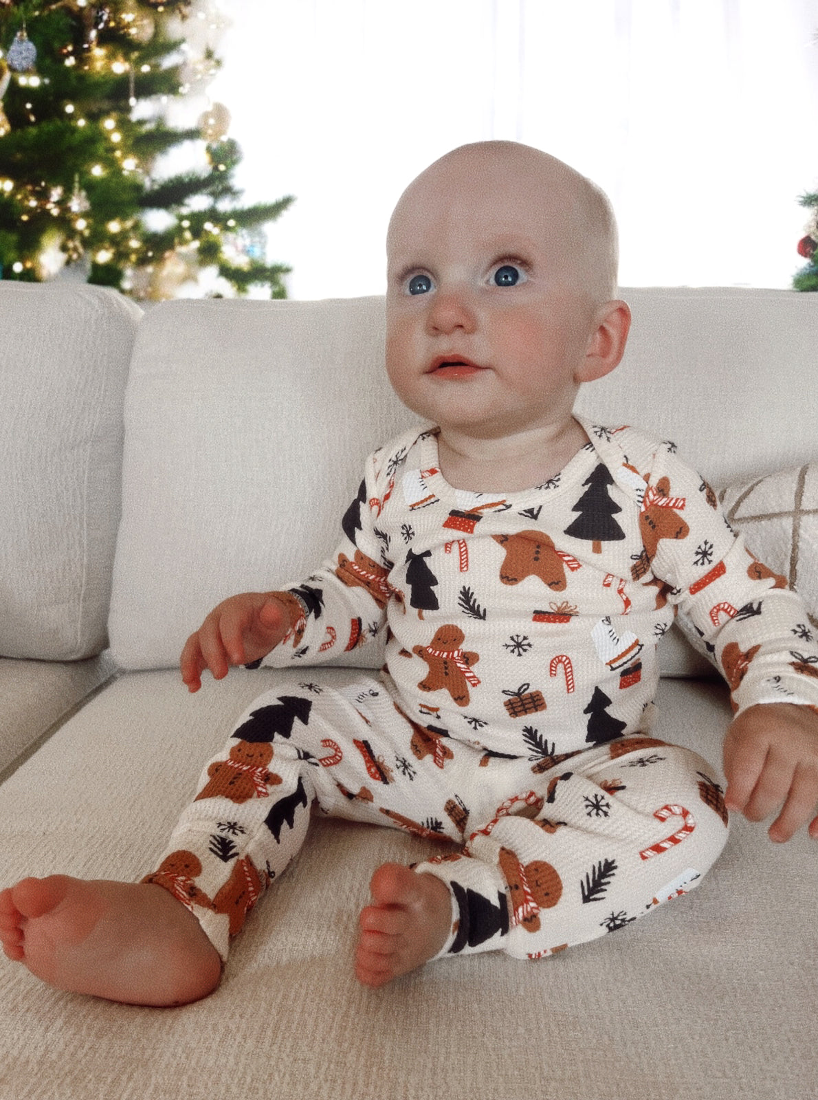 Baby in festive pajamas sits on a couch, looking up, with a decorated Christmas tree in the background.