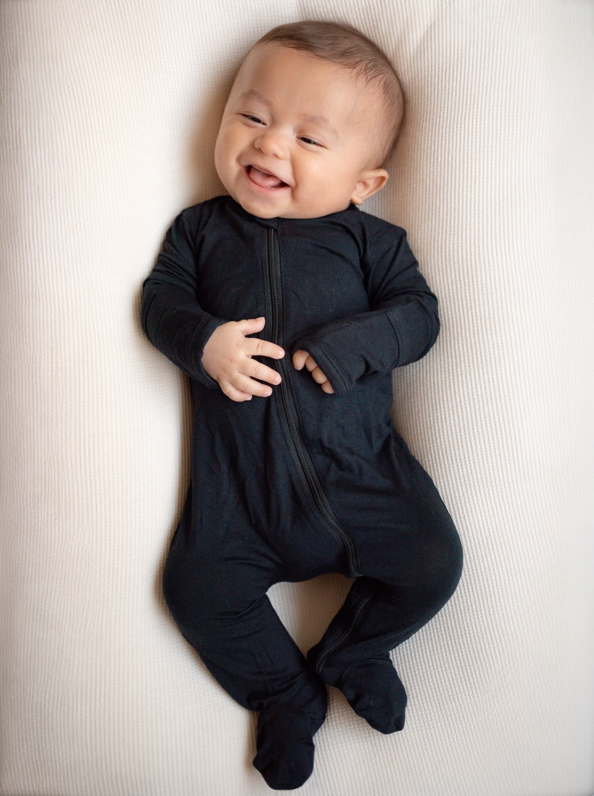 Smiling baby dressed in a black onesie, lying on a light textured surface.