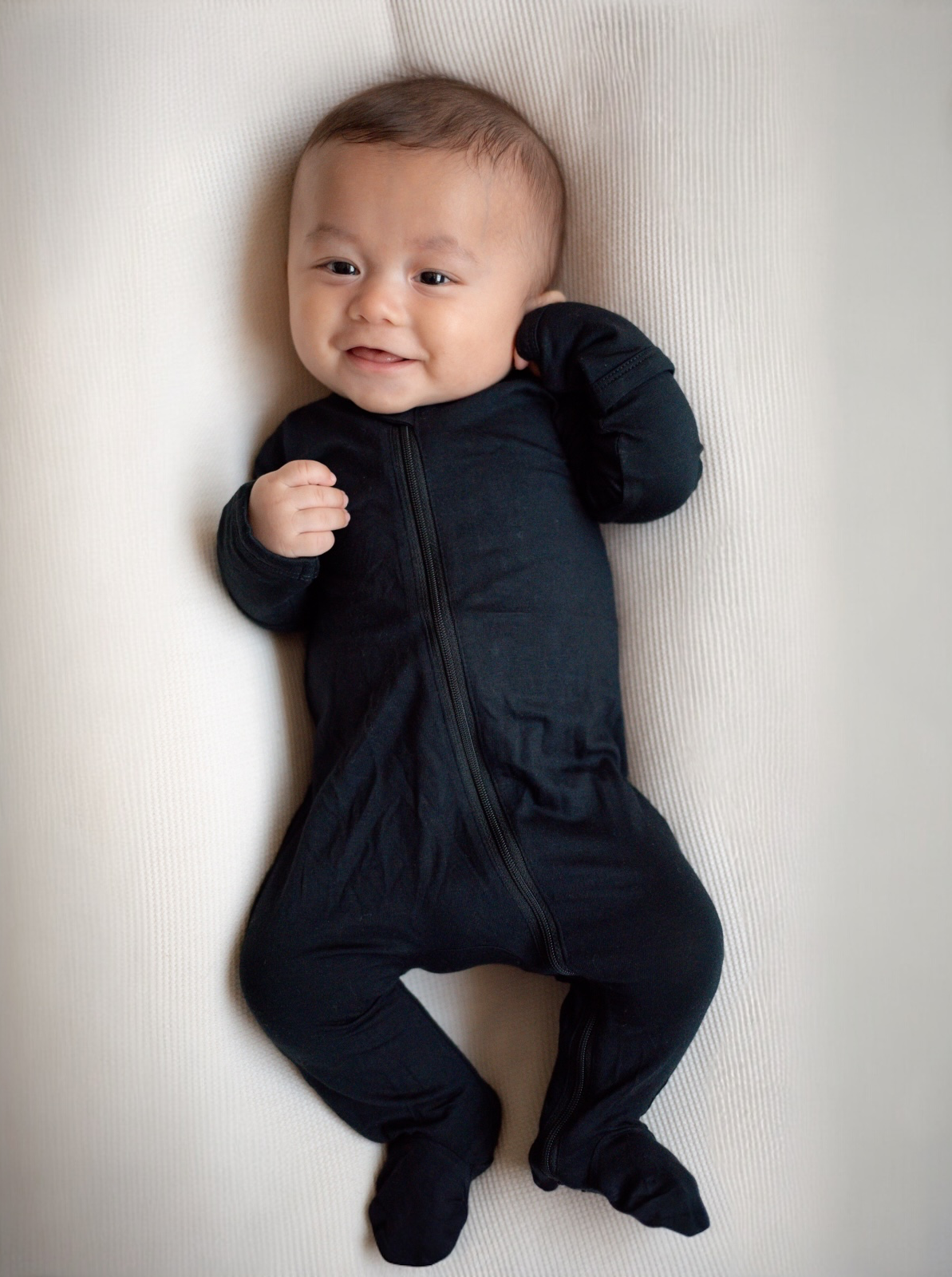 Smiling baby in a black onesie lying on a soft surface, hands near face, joyful expression.