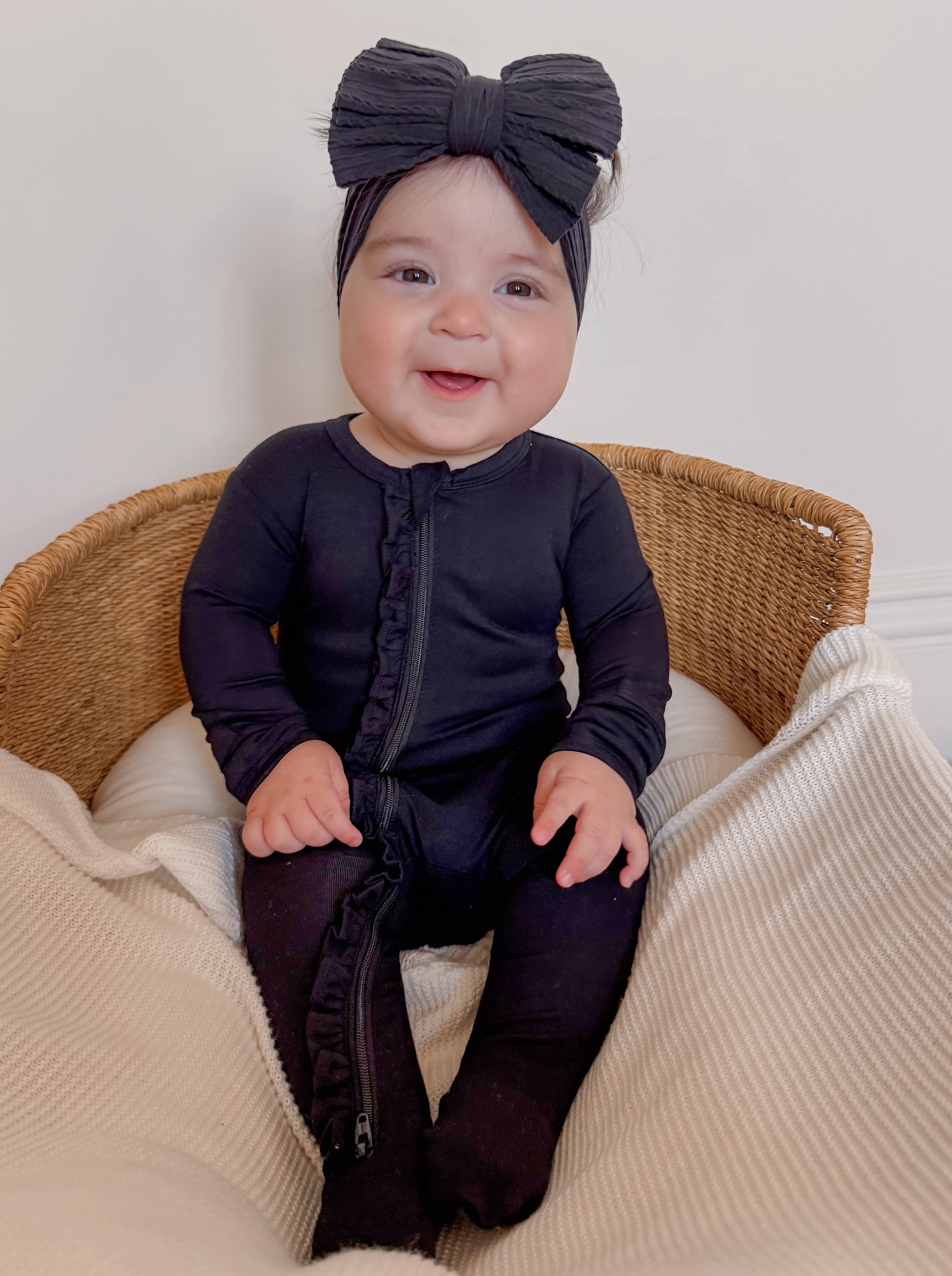 Smiling baby in a black outfit and headband, sitting on a blanket in a woven basket.