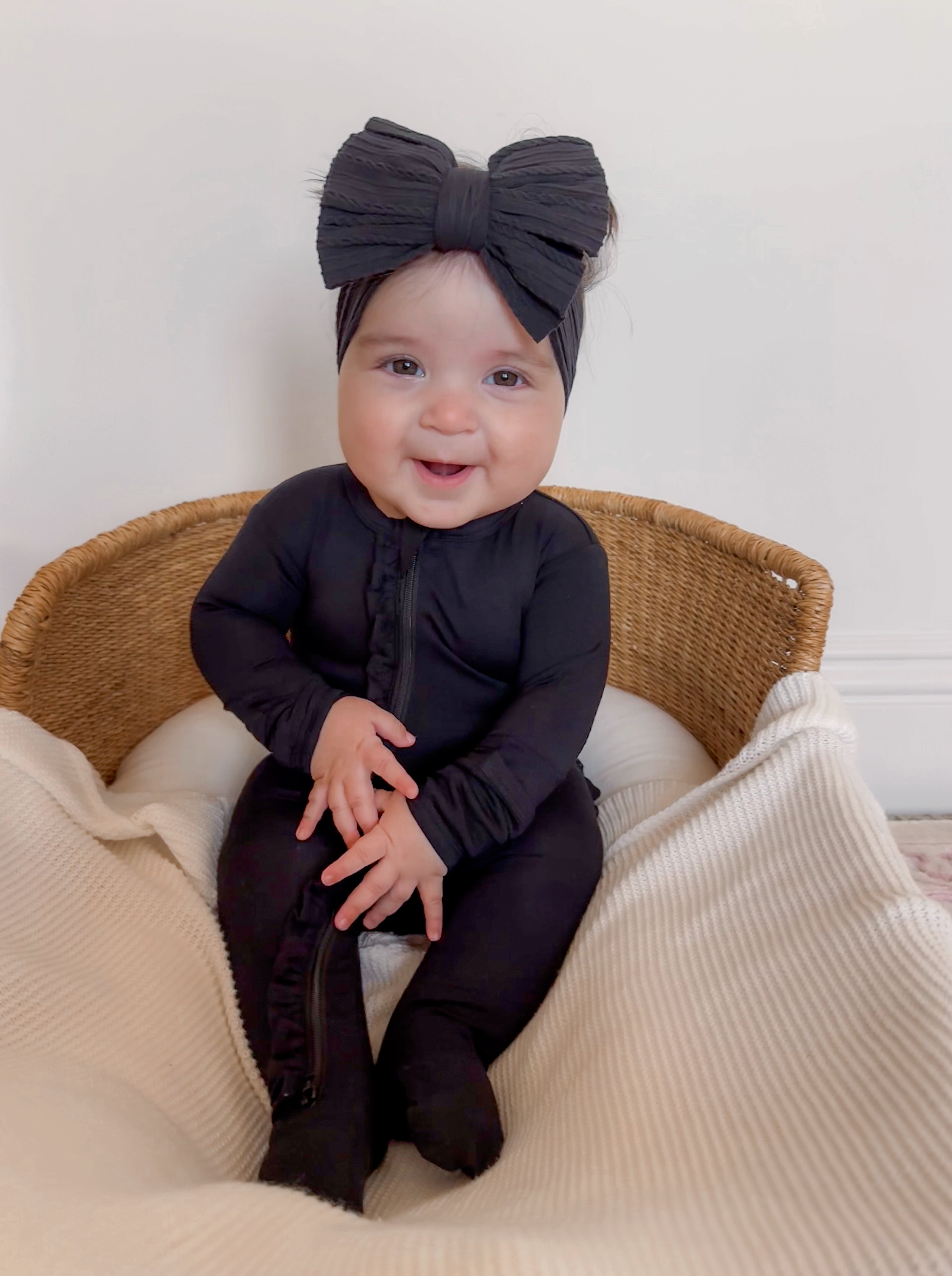 Smiling baby in black outfit and bow, sitting in a wicker chair on a soft blanket.