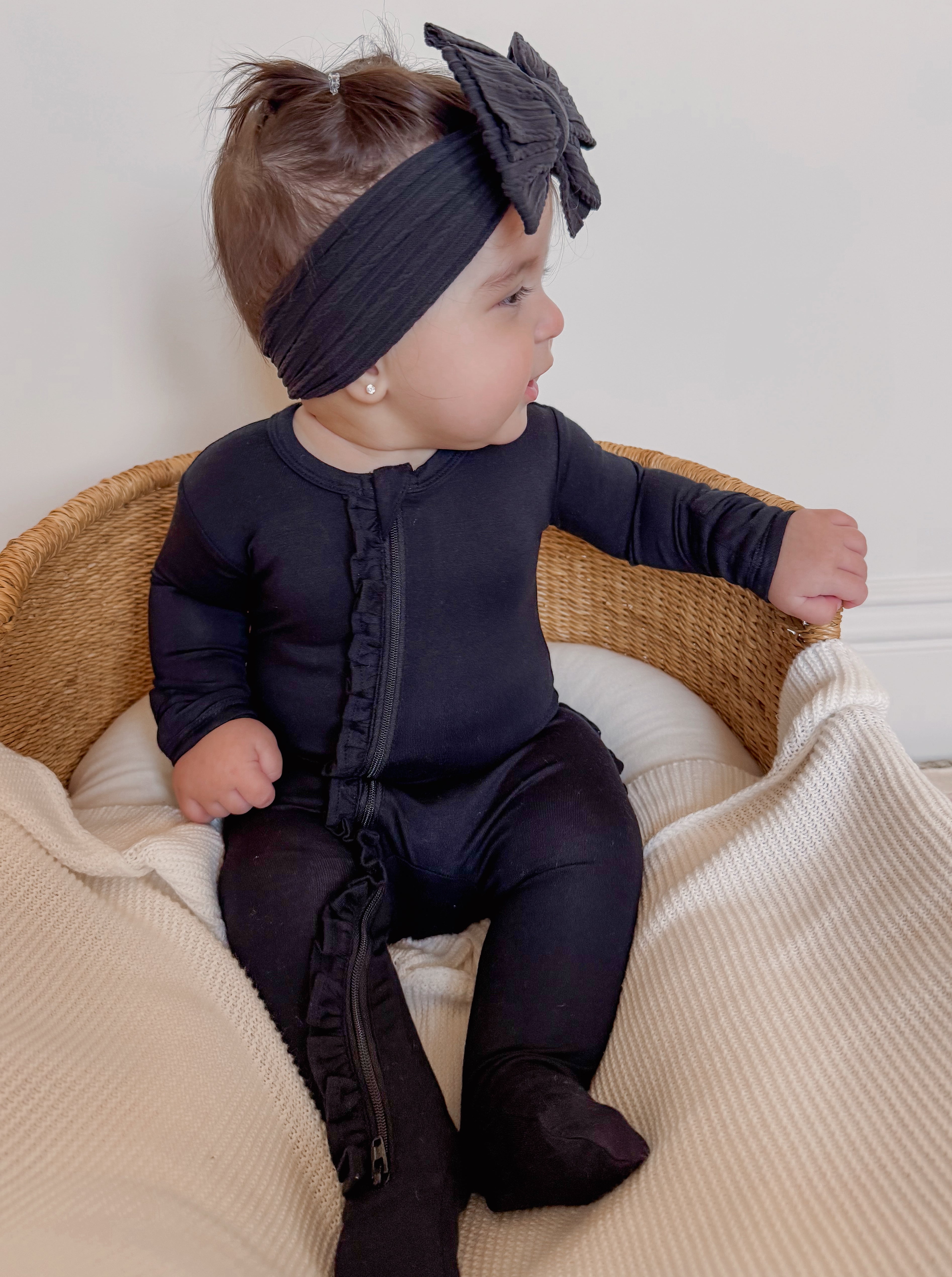 Baby in a black outfit with a ruffled zipper and a bow headband, sitting in a woven basket on a cozy blanket.