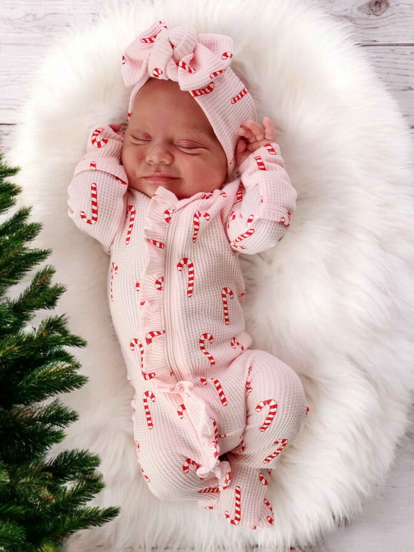Baby girl in a candy cane-patterned onesie and bow, peacefully sleeping on a fluffy white blanket.