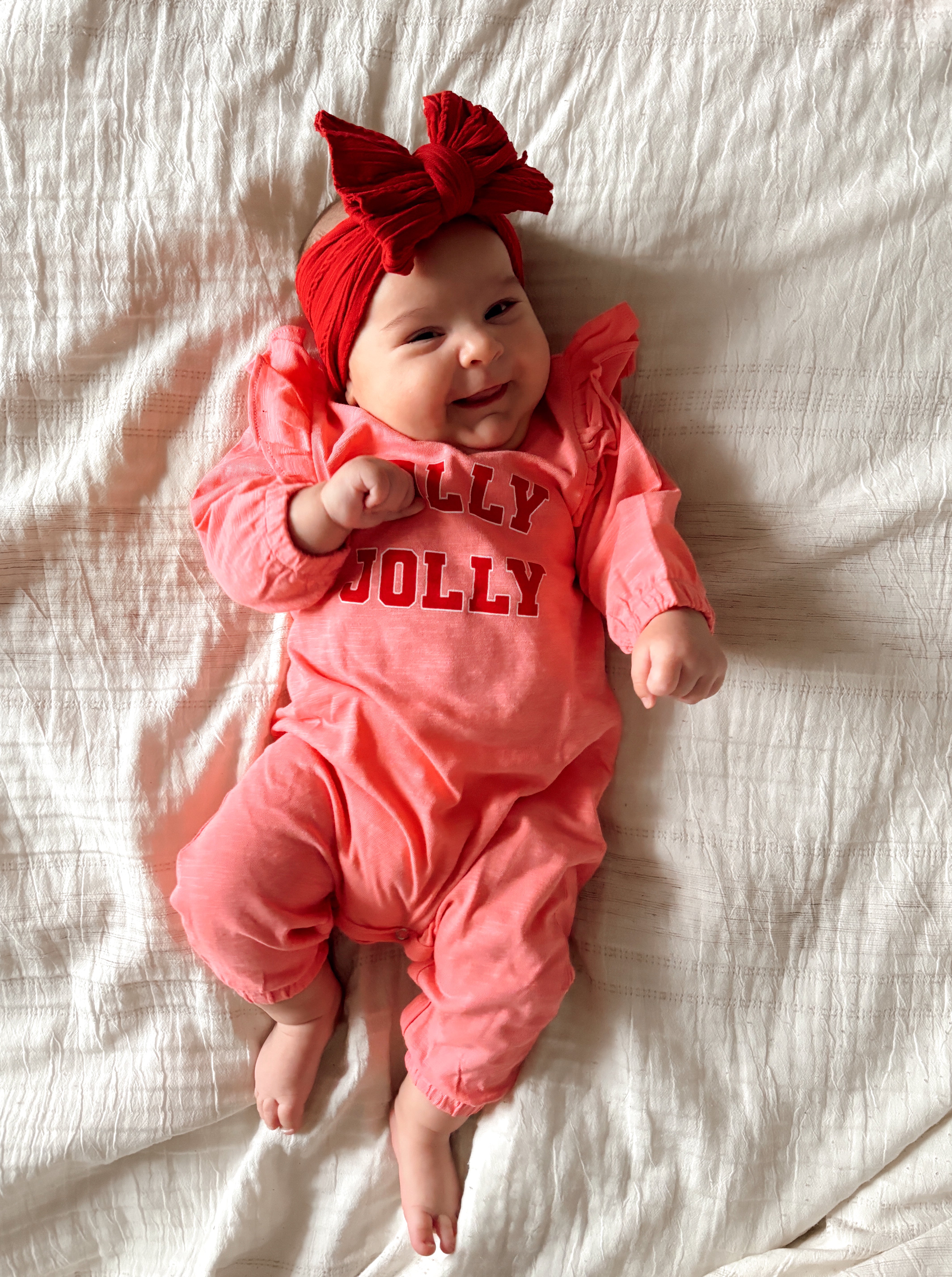 Smiling baby in a pink "Jolly Jolly" outfit with a red bow, lying on a white blanket.