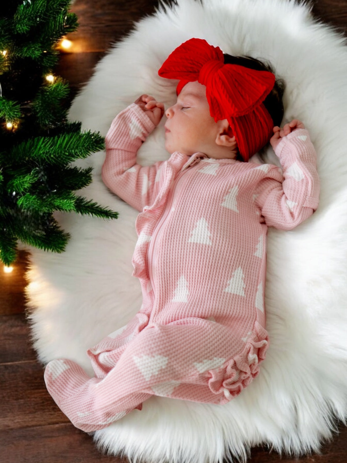 Infant girl in pink pajamas with white trees sleeps on a fluffy white rug by a Christmas tree.