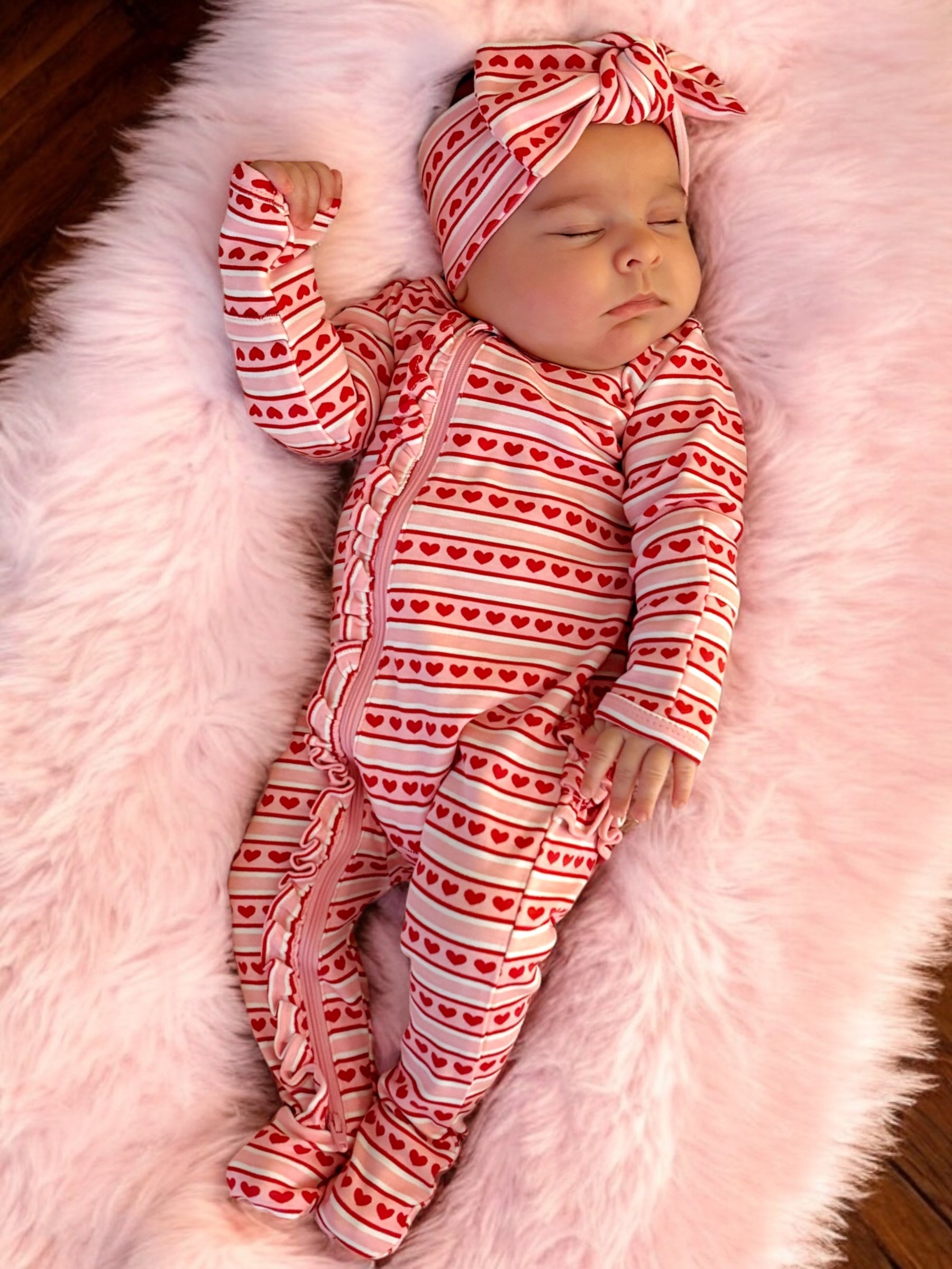 Baby girl in red and white heart-patterned pajamas and headband, peacefully sleeping on a fluffy pink blanket.