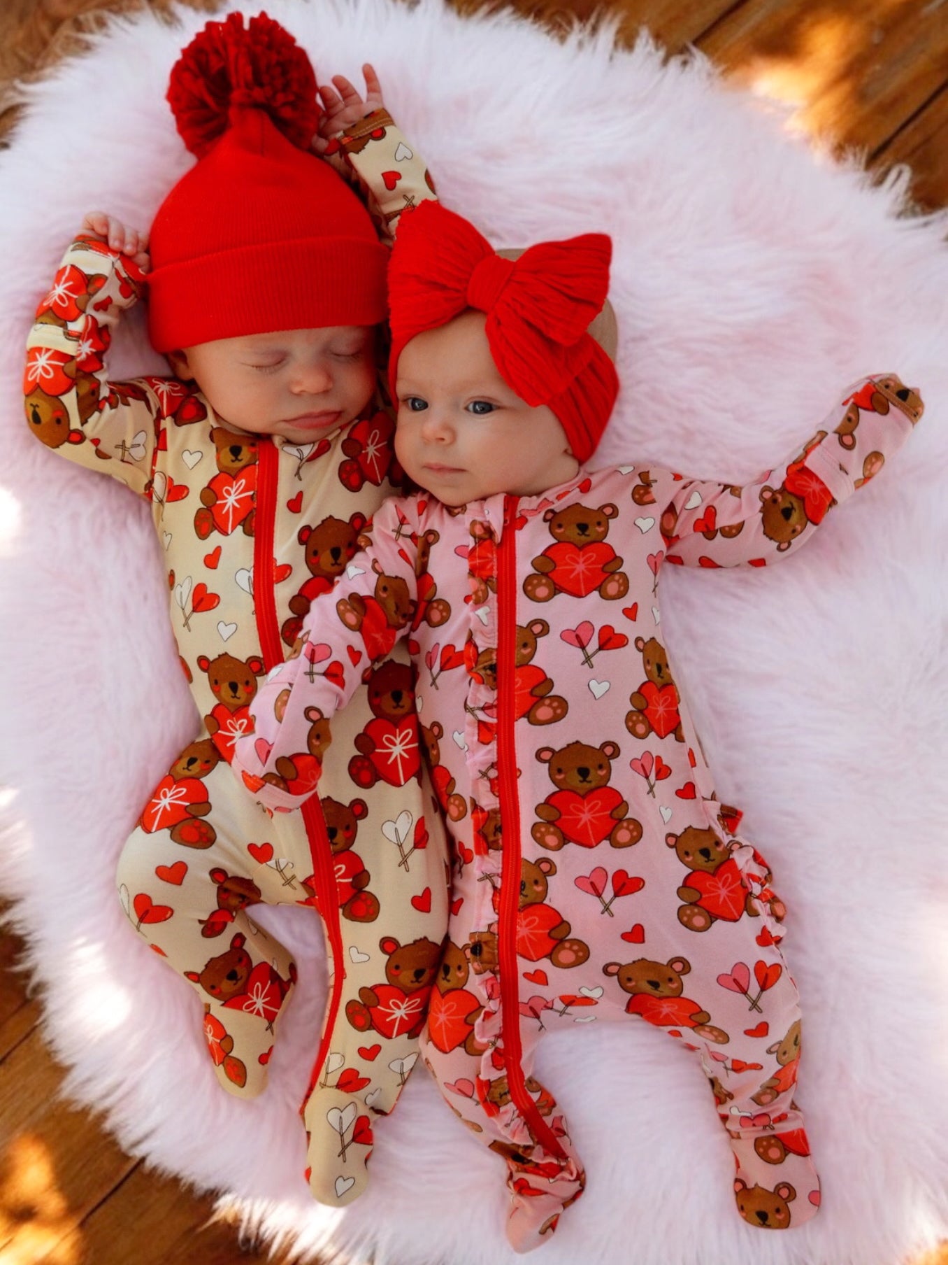 Two infants in colorful bear-patterned pajamas, one in a red hat and the other with a red bow, on a fluffy blanket.