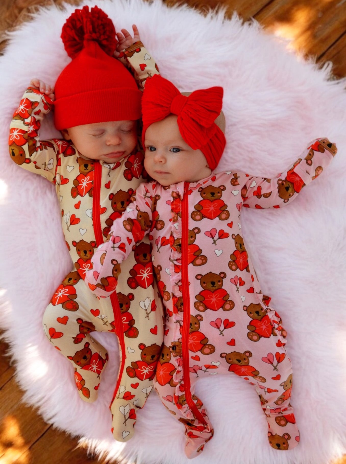 Two infants in colorful bear-patterned pajamas, one in a red hat and the other with a red bow, on a fluffy blanket.