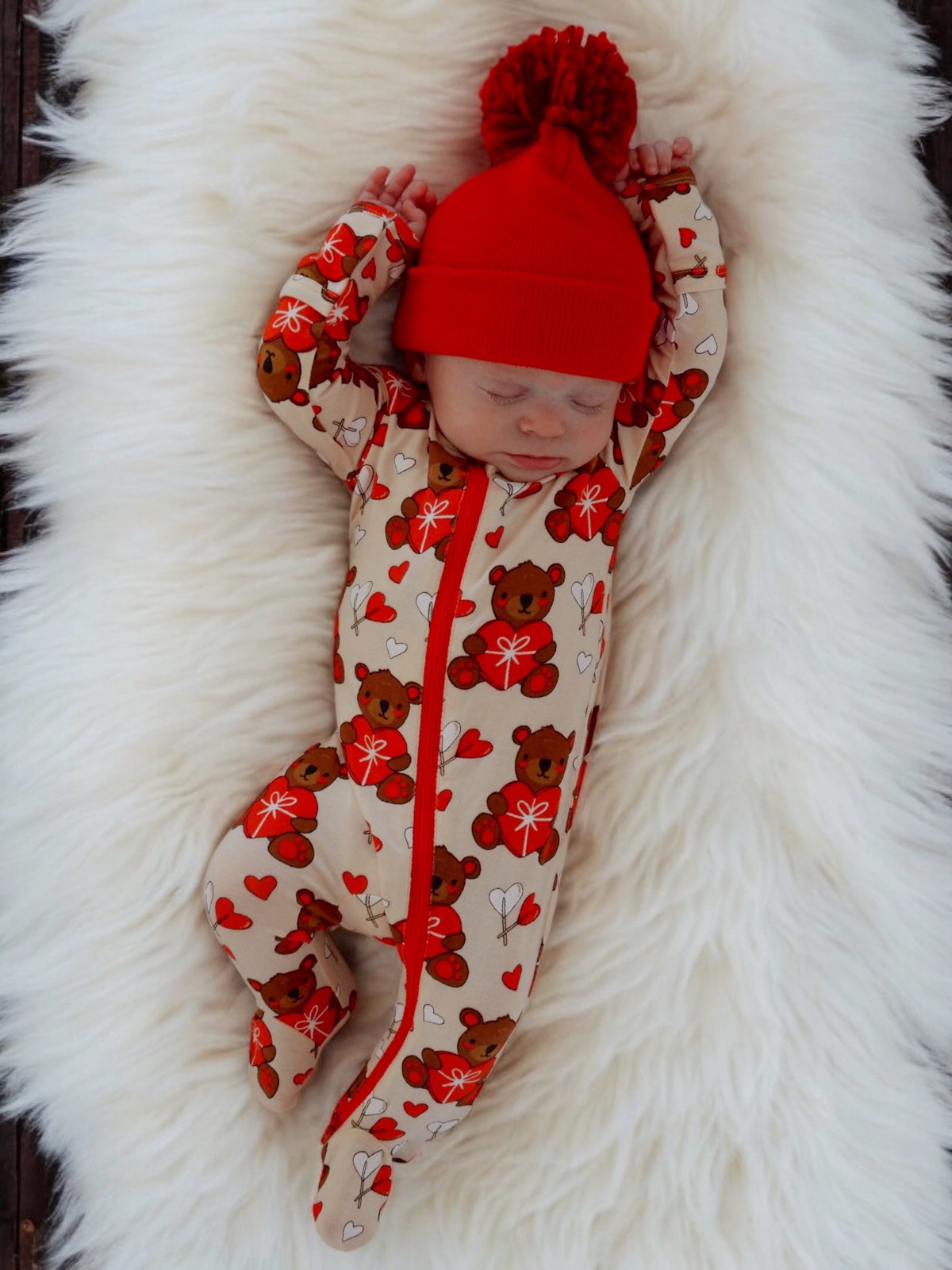 Infant sleeping on a fluffy white rug, wearing a red beanie and a bear-patterned onesie with hearts.
