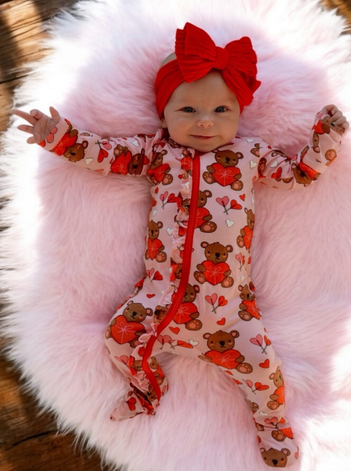 Smiling baby in a bear-themed onesie, wearing a red headband, lying on a fluffy pink blanket.