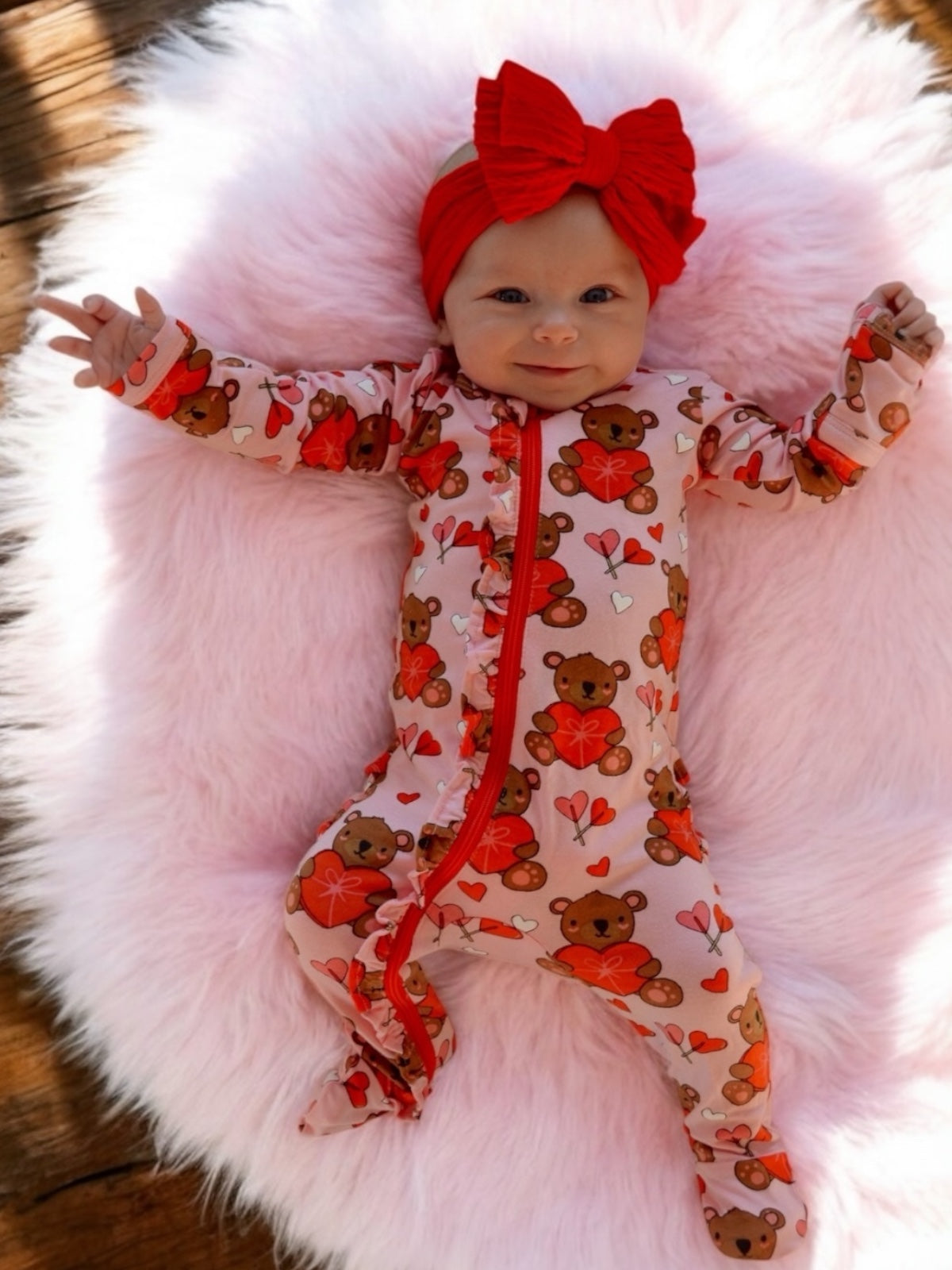 Smiling baby in a bear-themed onesie, wearing a red headband, lying on a fluffy pink blanket.