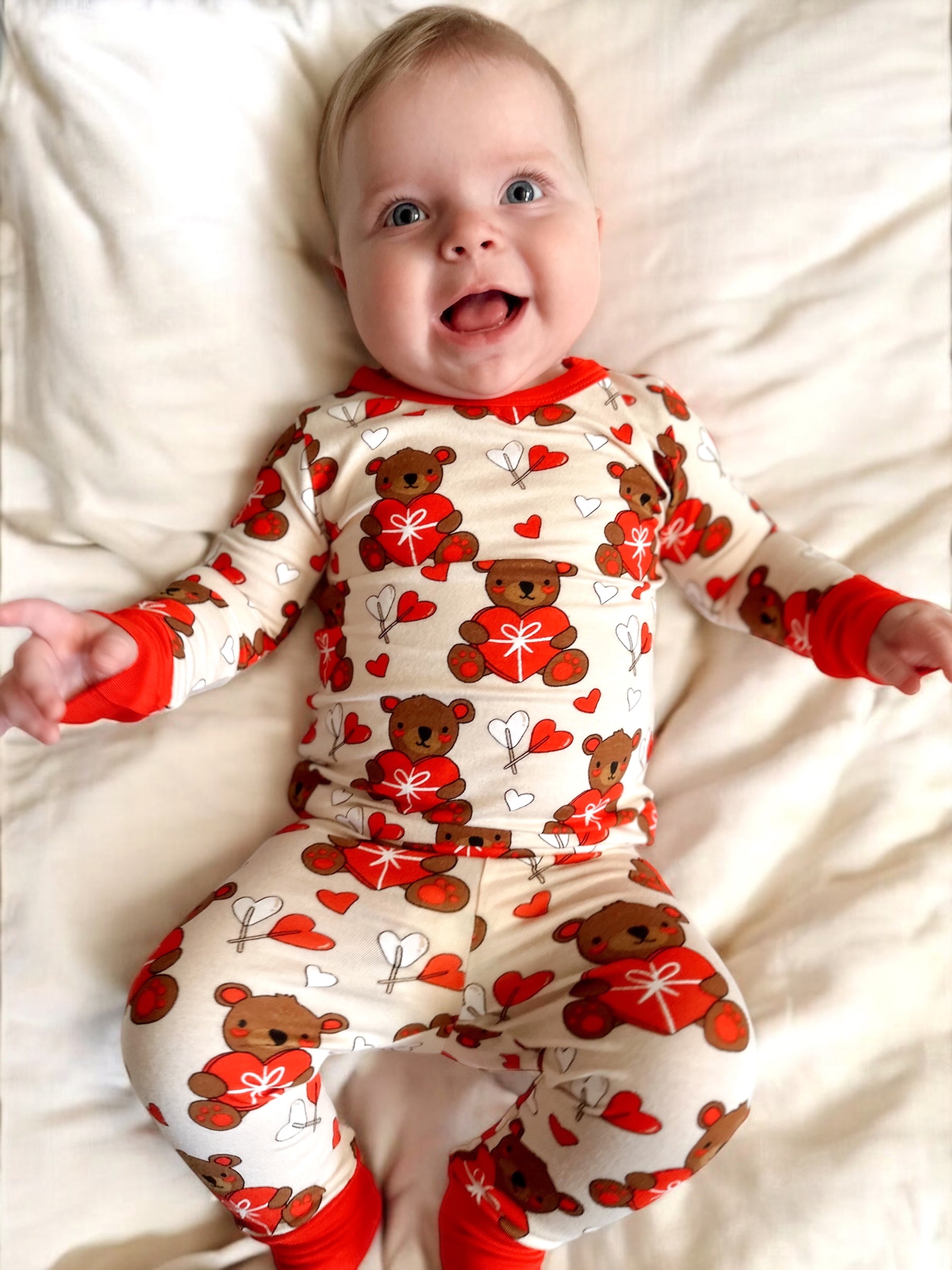 Smiling baby in bear-patterned pajamas, lying on a white blanket, with arms outstretched and joyful expression.