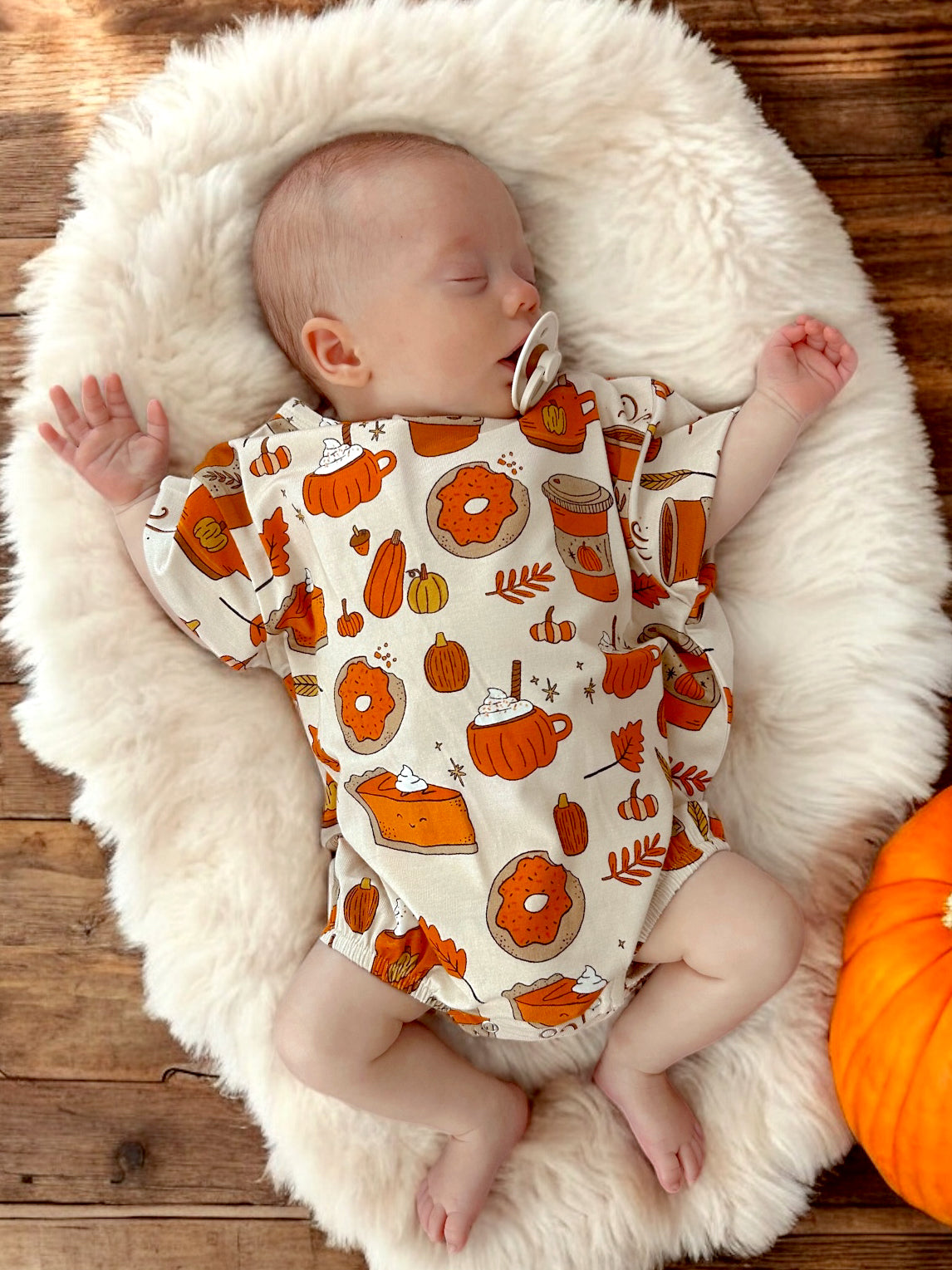 Sleeping baby on a fluffy blanket, wearing a shirt with autumn-themed prints like pumpkins and pies.