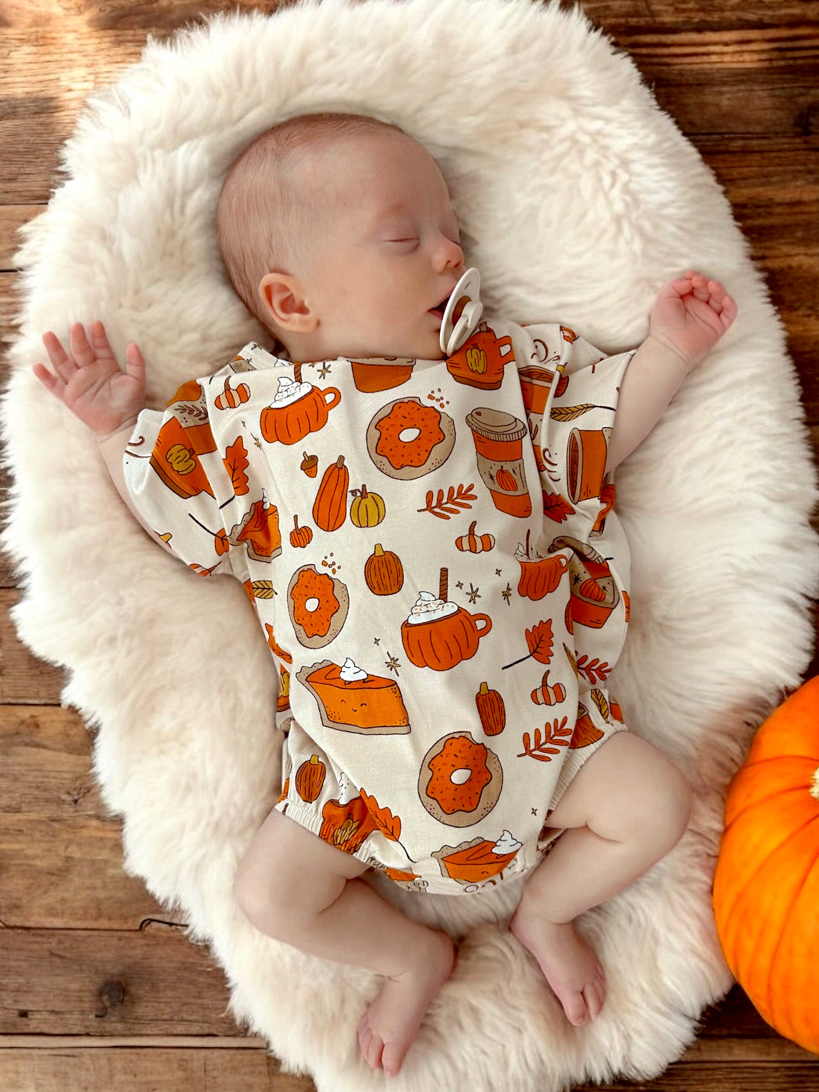 Sleeping baby on a fluffy blanket, wearing a shirt with autumn-themed prints like pumpkins and pies.