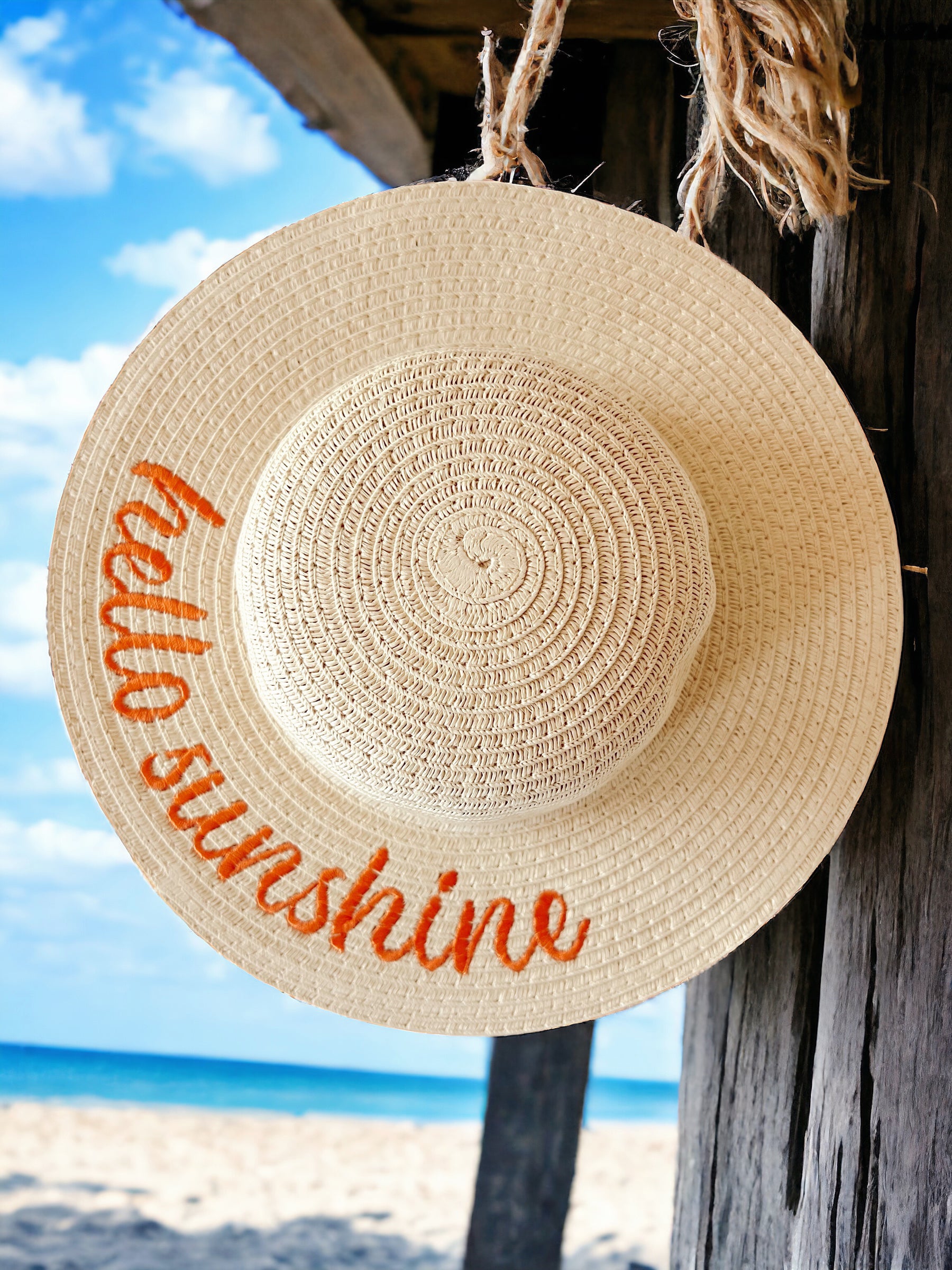 Straw hat hanging by the beach, with “hello sunshine” embroidered in orange on the brim, against a blue sky backdrop.