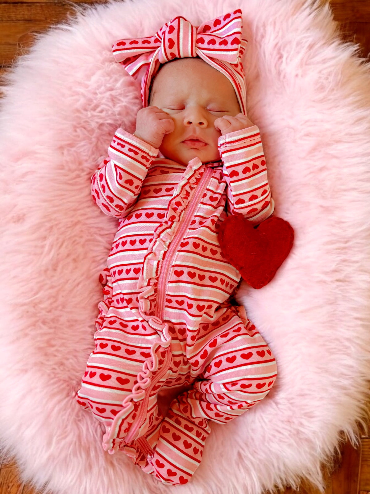 Newborn in a heart-patterned onesie and bow, resting on a pink fluffy blanket with a small red heart toy.