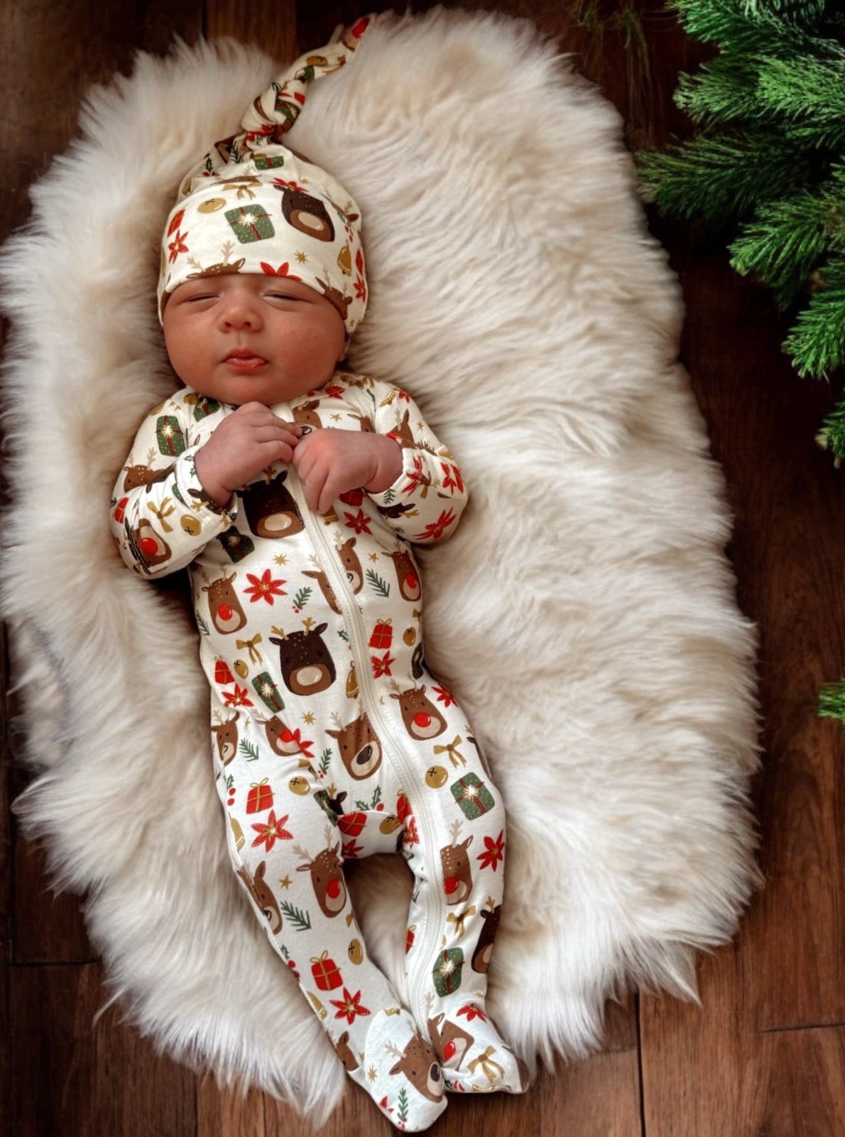 Baby wearing a festive animal-print onesie and hat, resting on a fluffy white rug with greenery nearby.