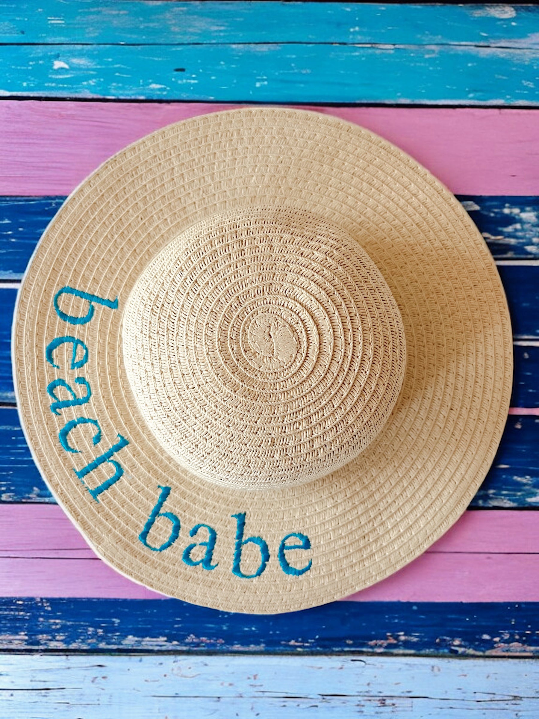Wide-brimmed straw hat with "beach babe" embroidered in turquoise on a colorful wooden background.