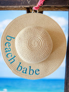 Woven straw hat with "beach babe" embroidered, hanging against a blue ocean backdrop.